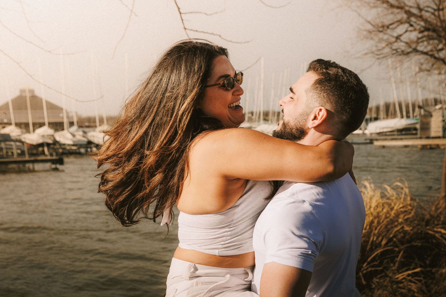 Couple in white clothing embraces passionately during a waterfront photo session at golden hour.