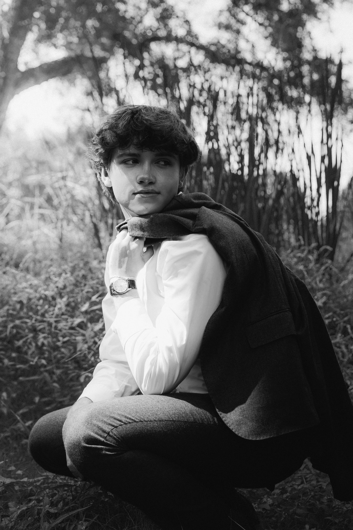 Black and white portrait of someone in a white sweater and dark vest crouching outdoors among trees.