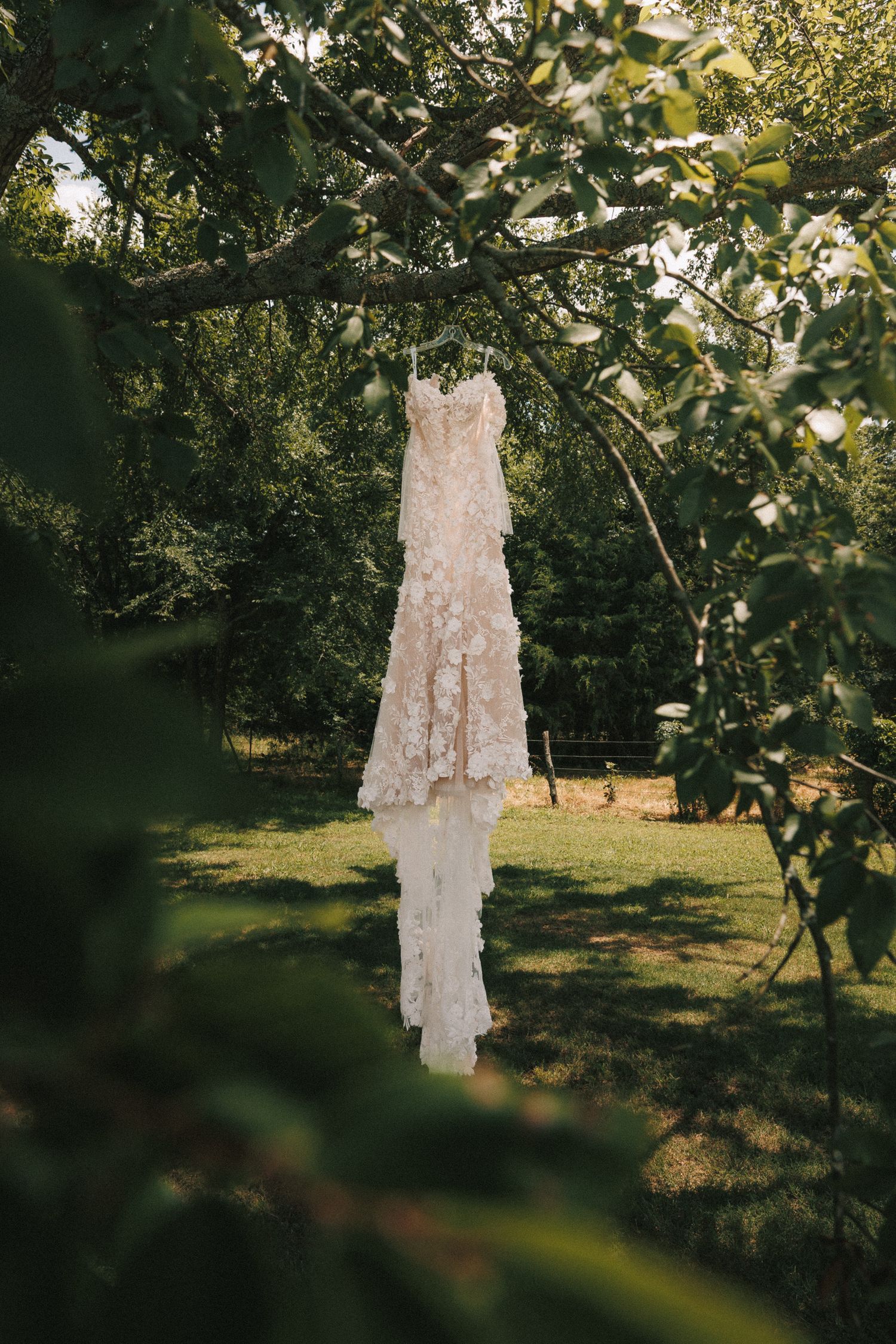 An ornate white chandelier hanging from tree branches in a sunlit garden setting.