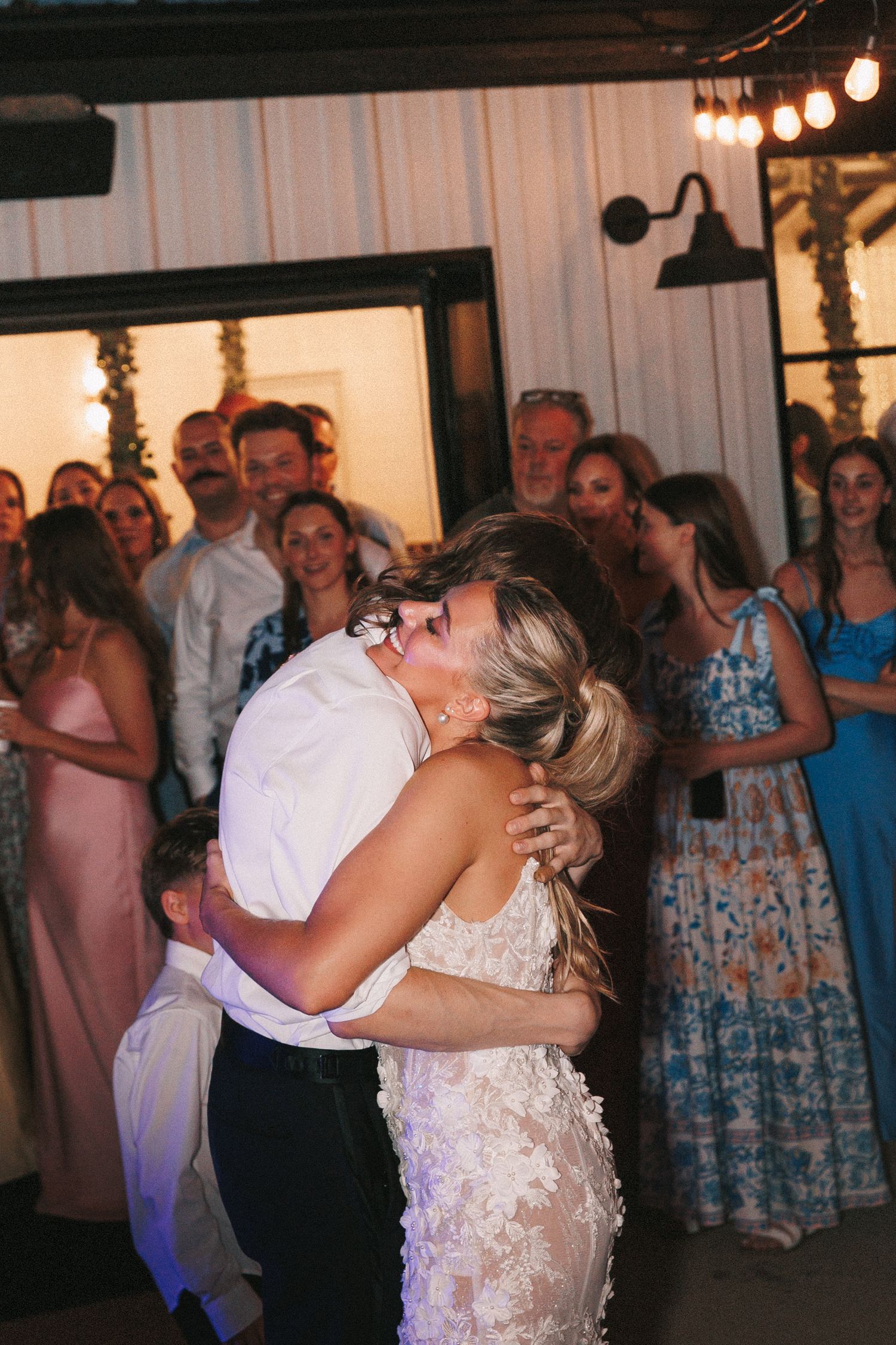 Wedding party dancing and celebrating on a reception dance floor beneath hanging lights and white drapery.