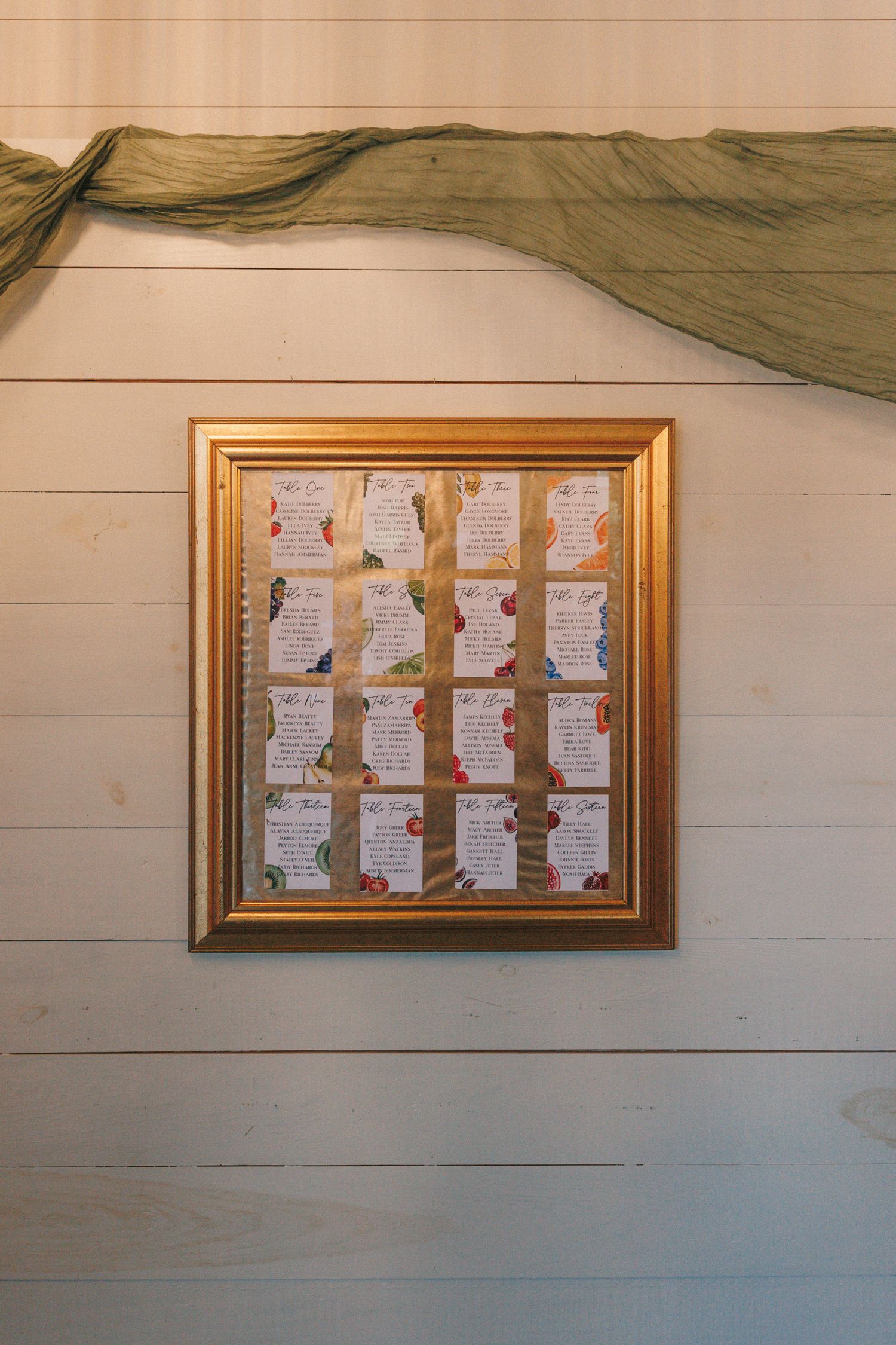 Gold-framed seating chart display mounted on white shiplap wall with flowing fabric draping above.