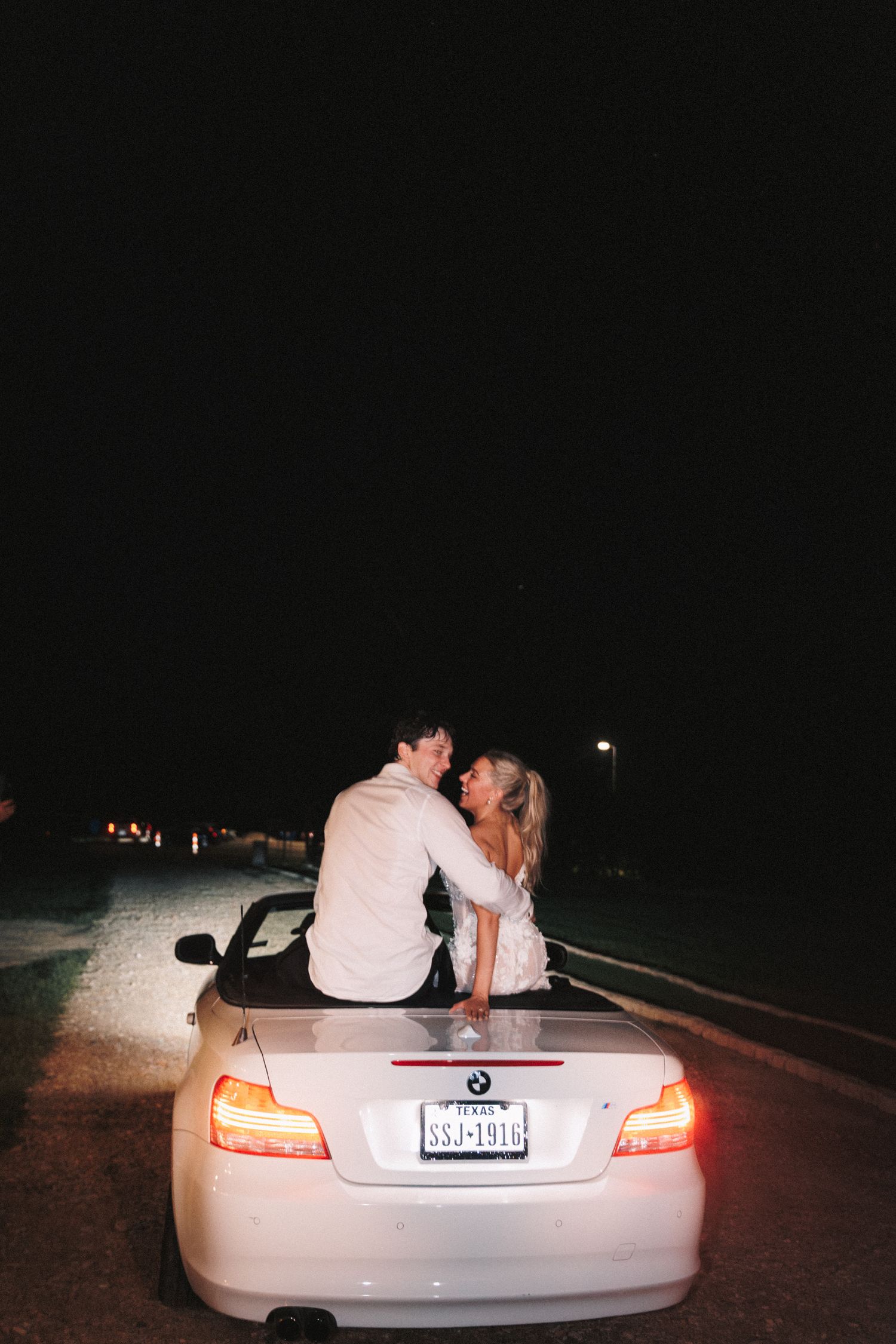 Bride and groom sitting romantically on the trunk of their getaway car with taillights glowing.