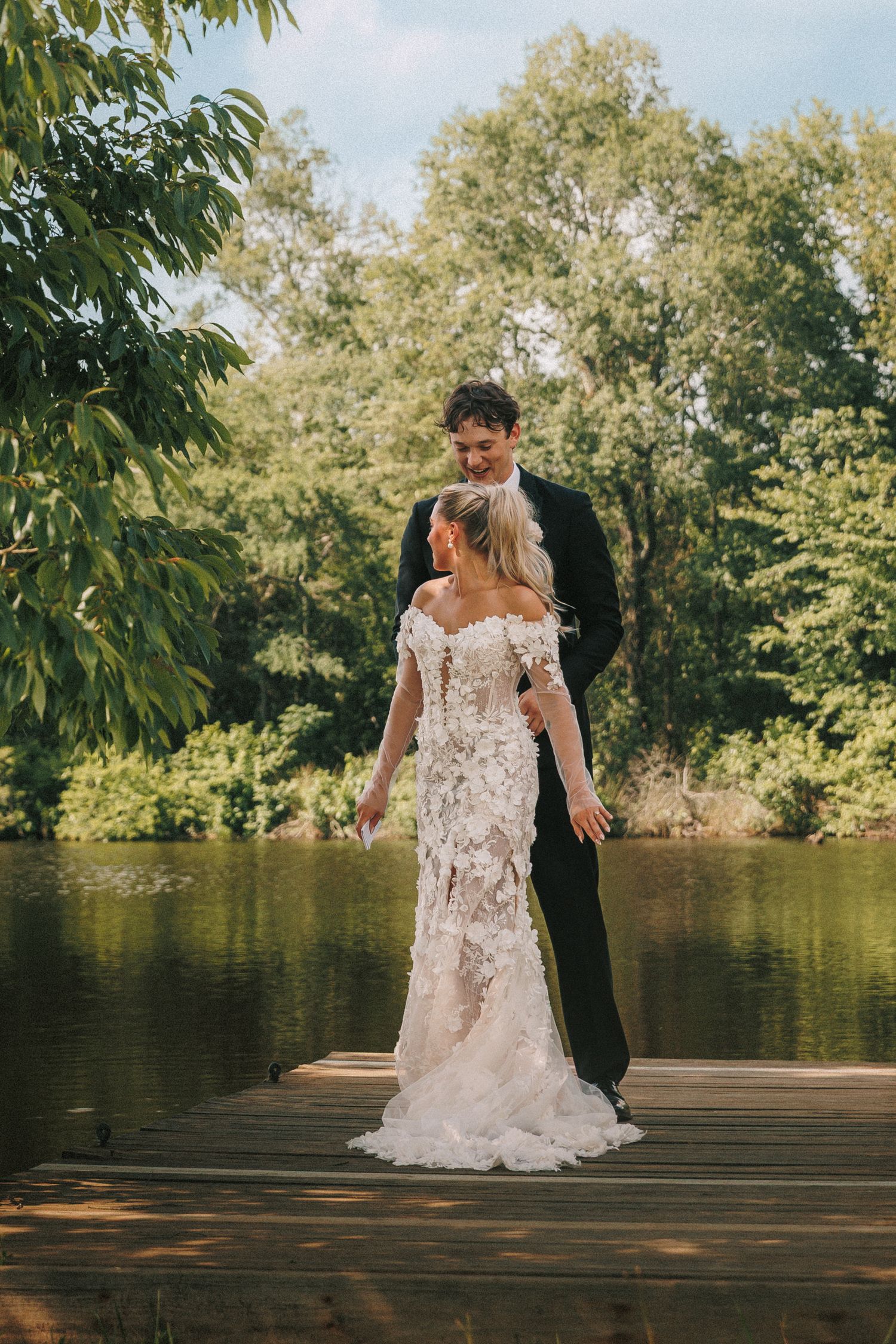 A romantic wedding couple poses together on a wooden dock by a serene lake surrounded by lush green trees.