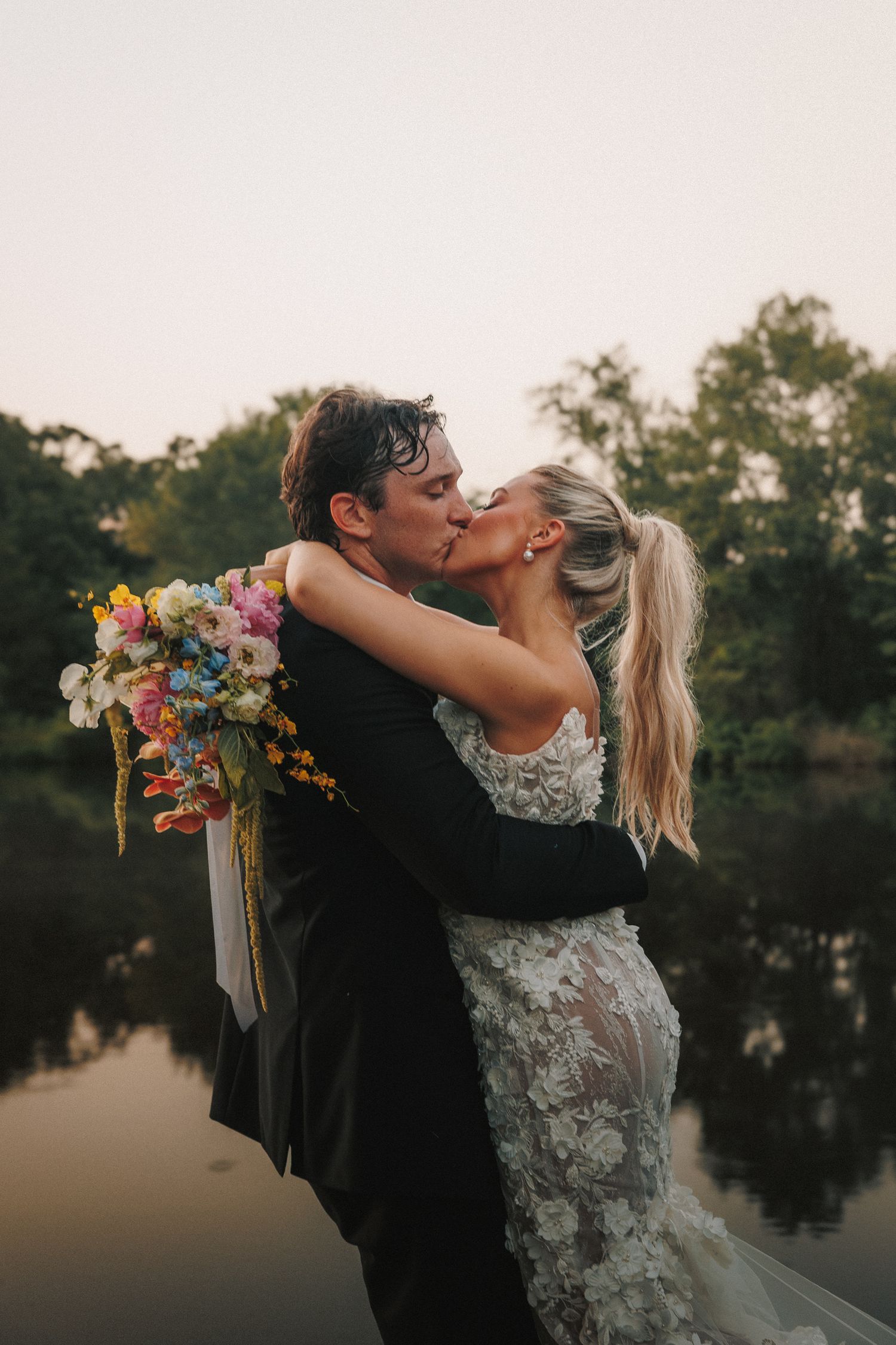A couple in wedding attire share romantic kisses at sunset by a serene lake with trees in the background.