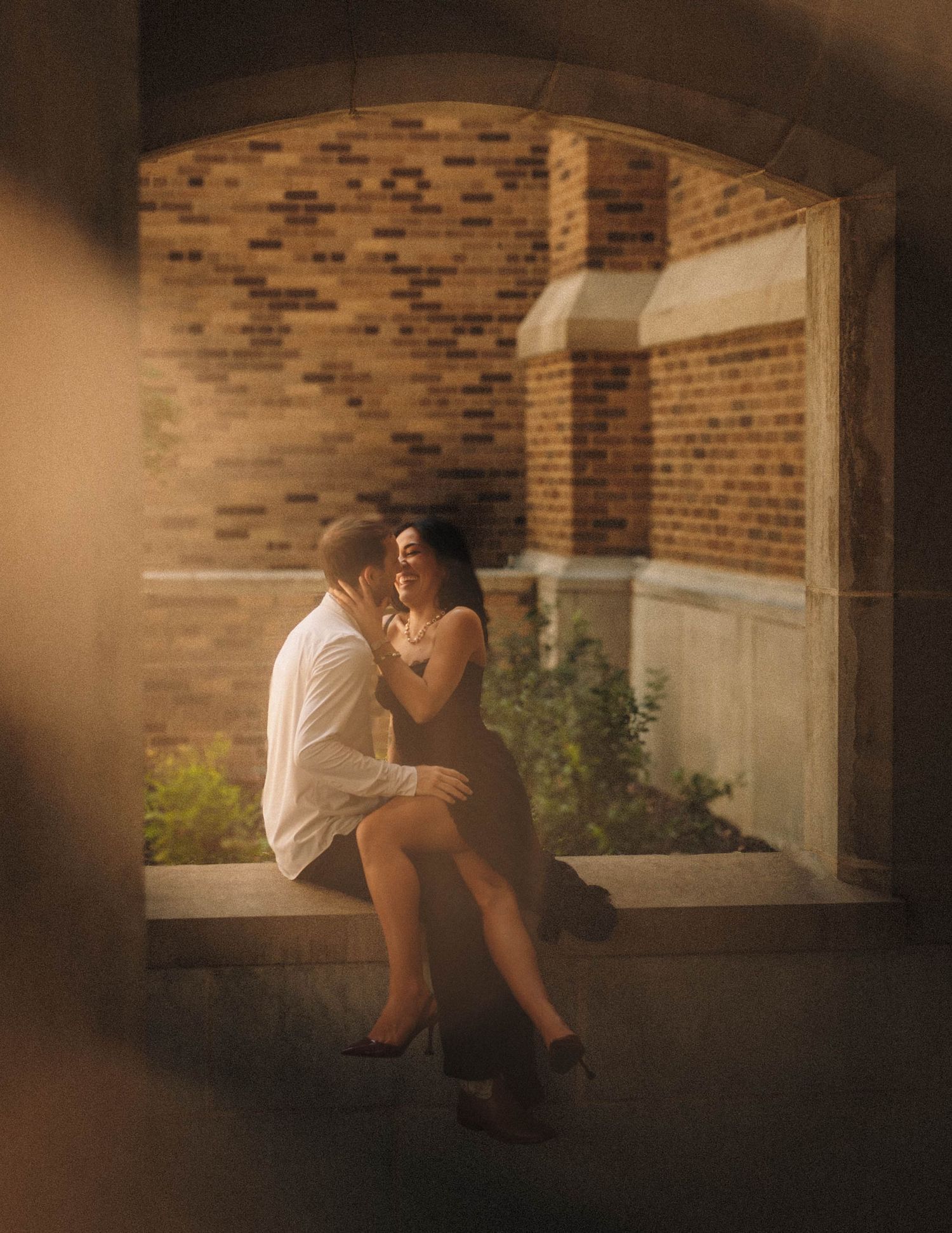 A romantic couple shares a quiet moment together on a bench against a brick wall in warm, moody lighting.
