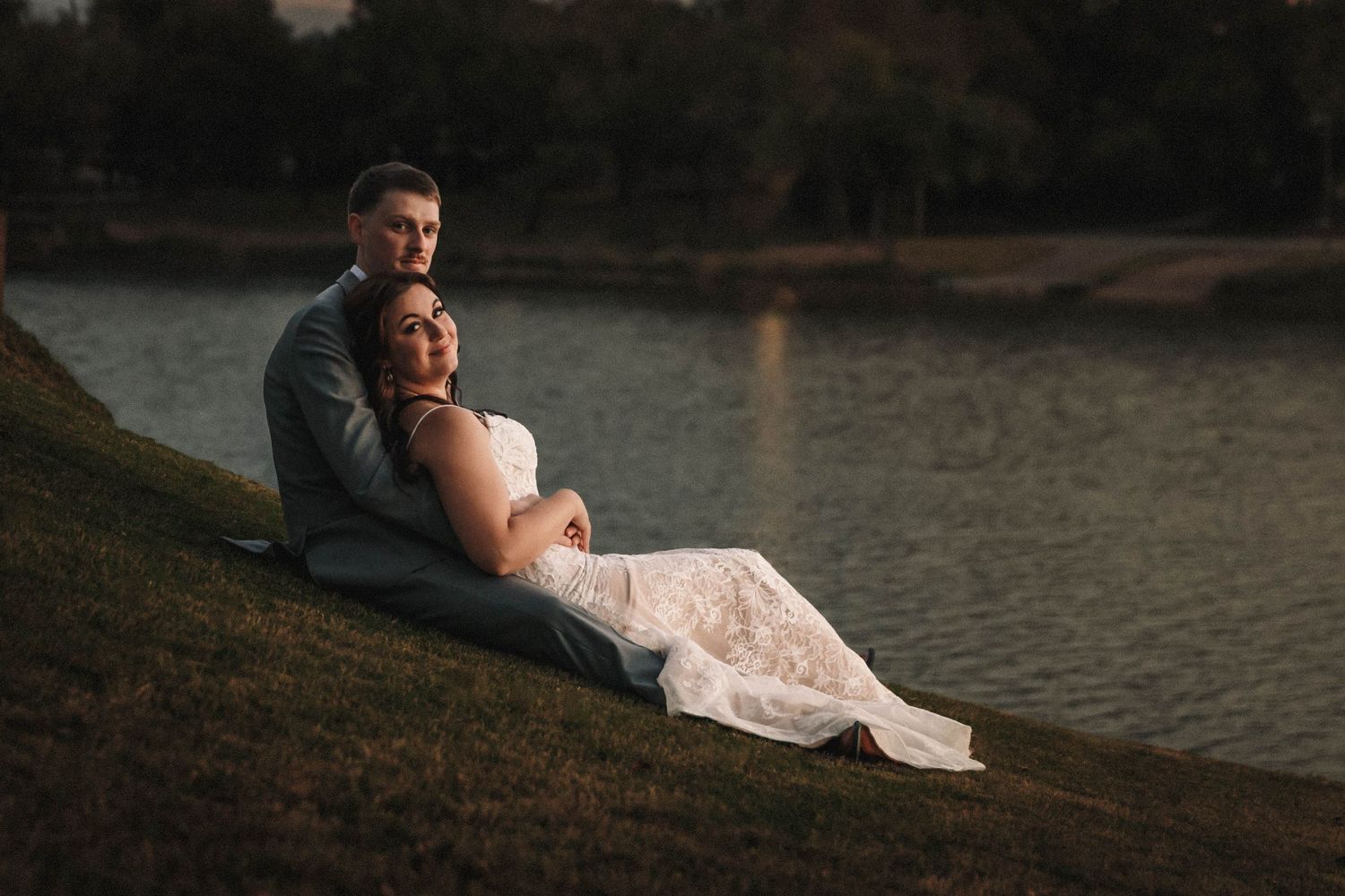 A couple in wedding attire sits intimately by a waterfront during a romantic sunset photo session.
