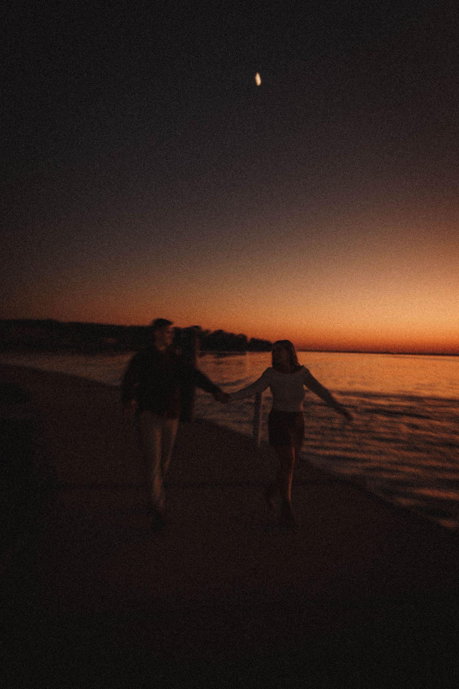 Silhouettes walk along a beach at sunset with orange sky reflecting on the water.