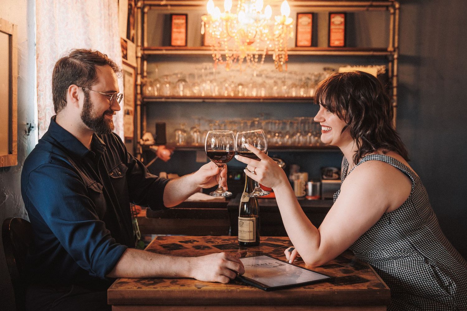 Two people sit together examining menus at a dimly lit restaurant table with wine glasses.