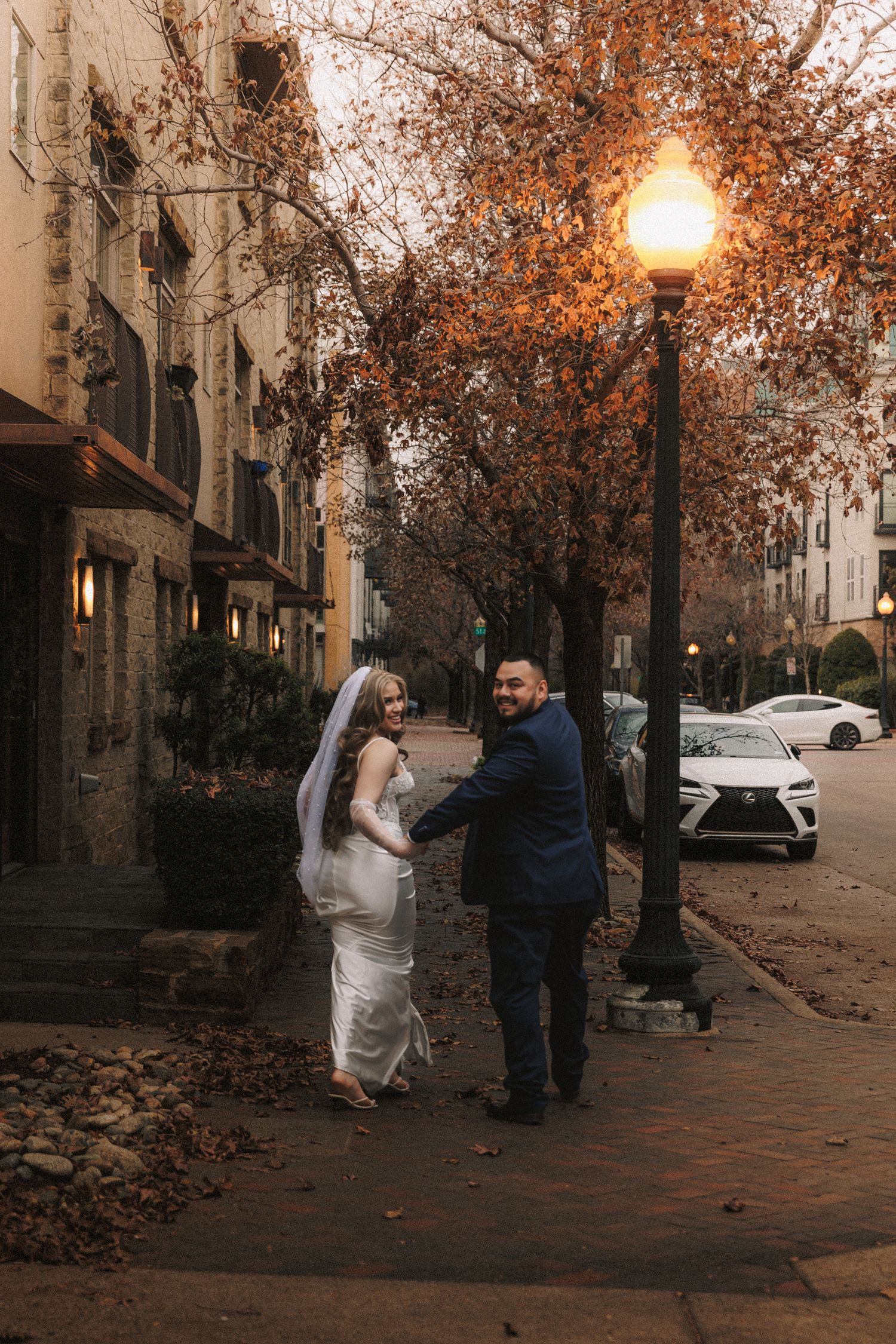 A couple walks along a brick sidewalk on an autumn evening with glowing street lamps and fallen leaves.