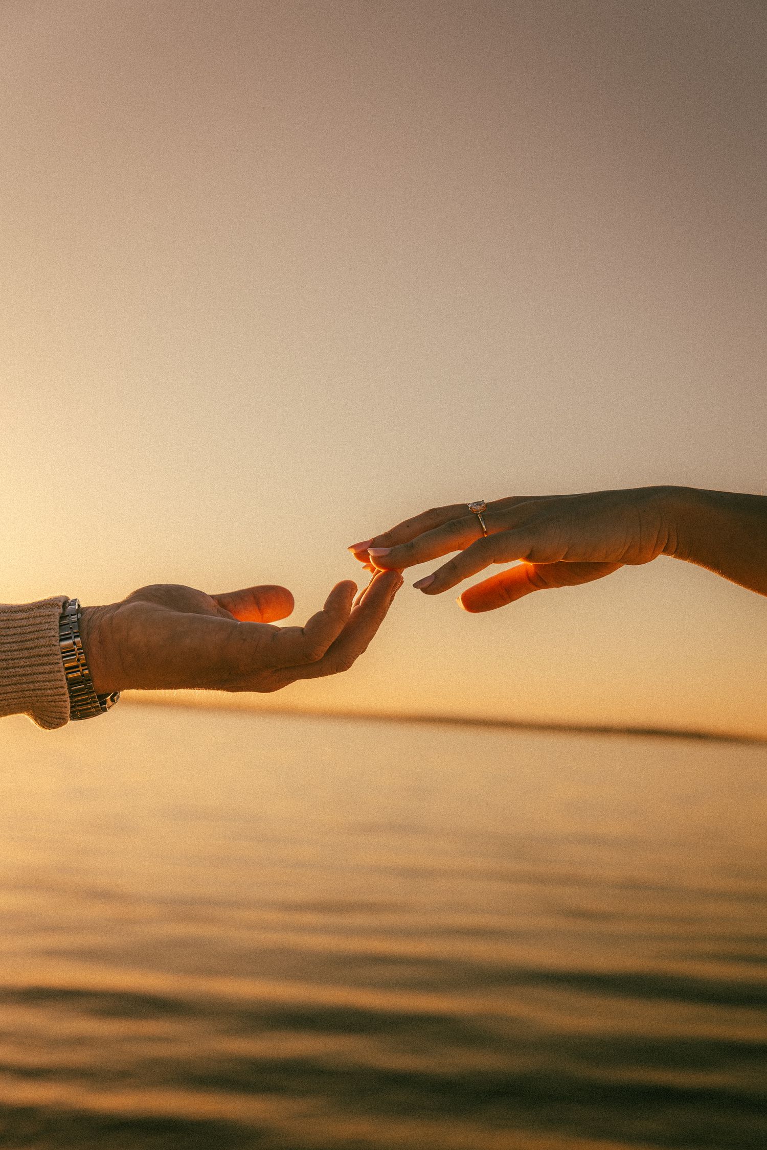 Two hands reaching towards each other against a golden sunset over calm water.