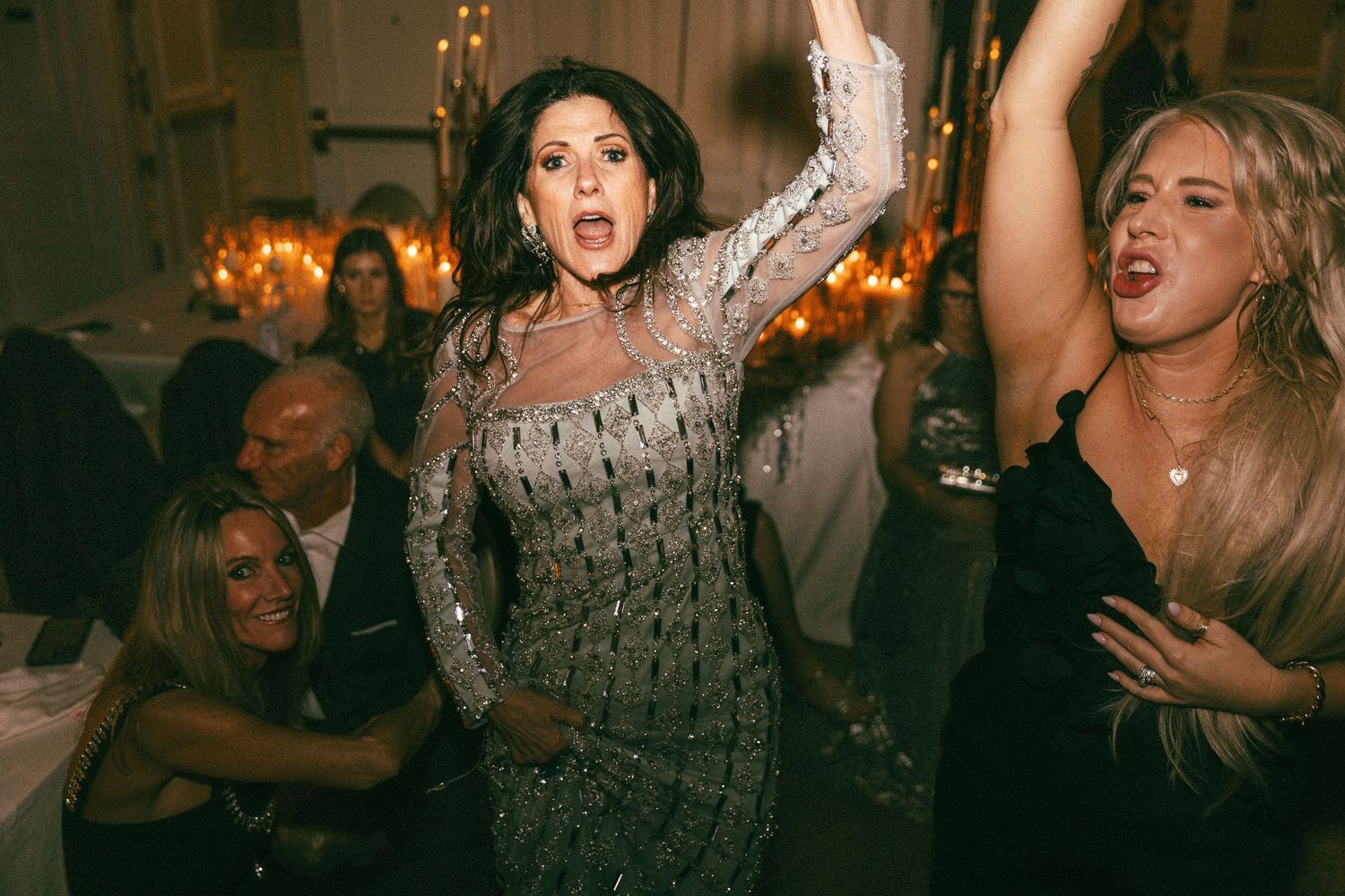 Wedding guest in silver beaded gown dances joyfully with arms raised at reception.