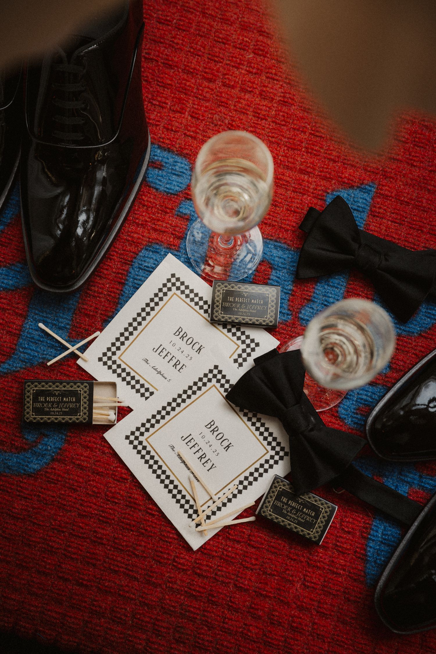 Wedding flat lay with black bow ties, dress shoes, invitation cards for Brock and Jeffrey, matchboxes, and champagne glasses on red textile.