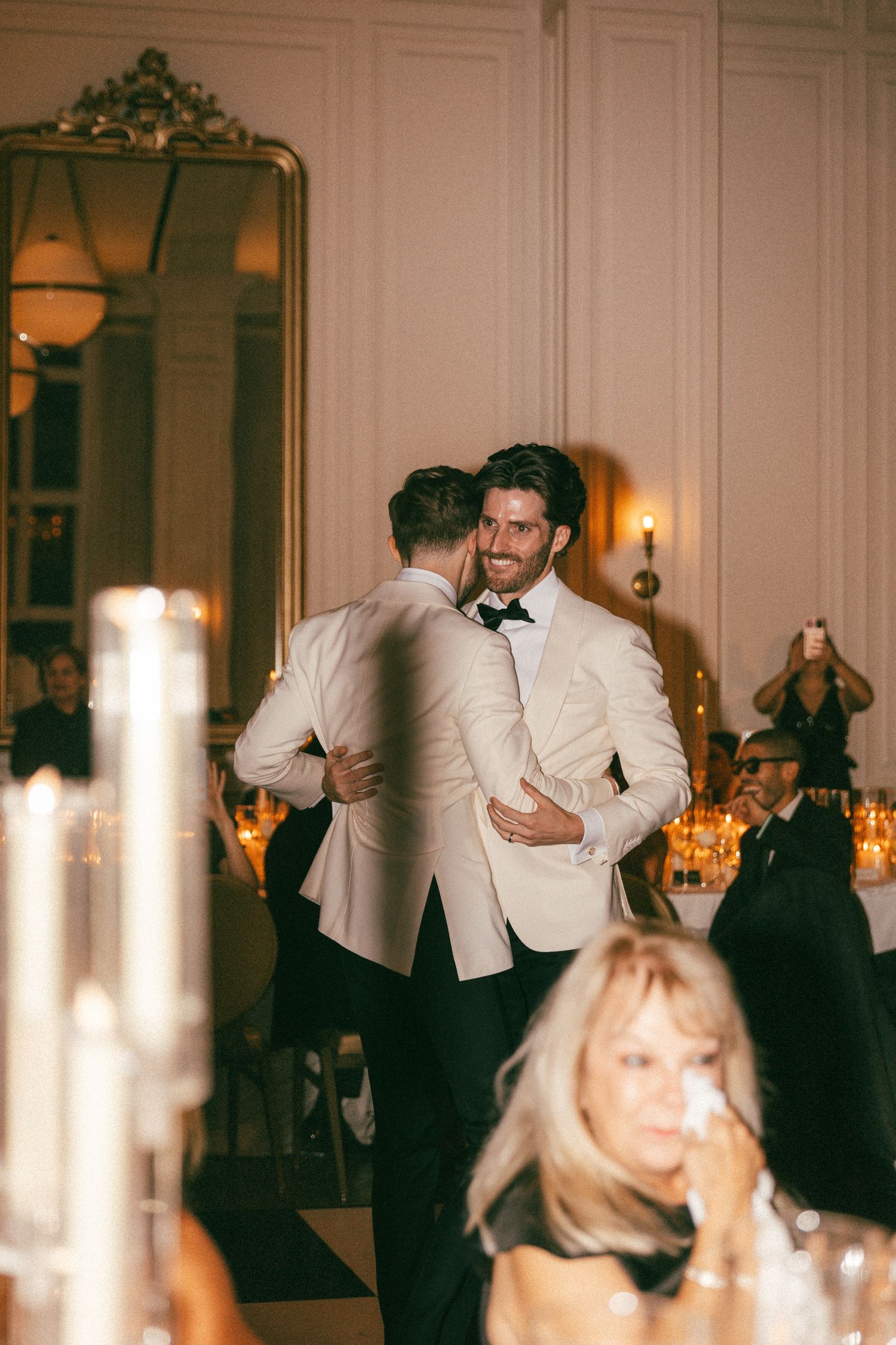 Two men in white tuxedo jackets embracing warmly on wedding reception dance floor surrounded by candlelight.