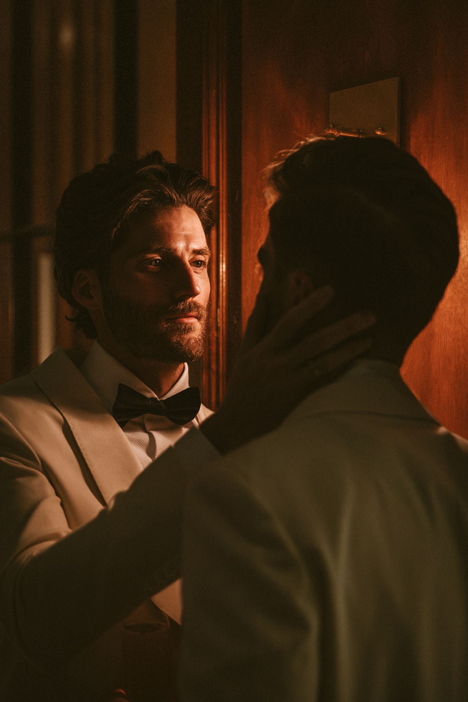 Two grooms in white tuxedos with bow ties sharing an intimate moment in warm golden light.