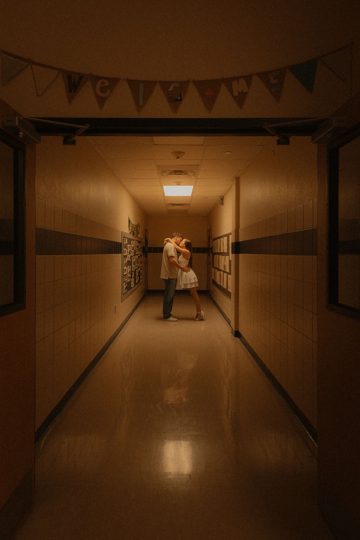 Couple sharing a romantic kiss in a dimly lit school hallway with bunting banner overhead.