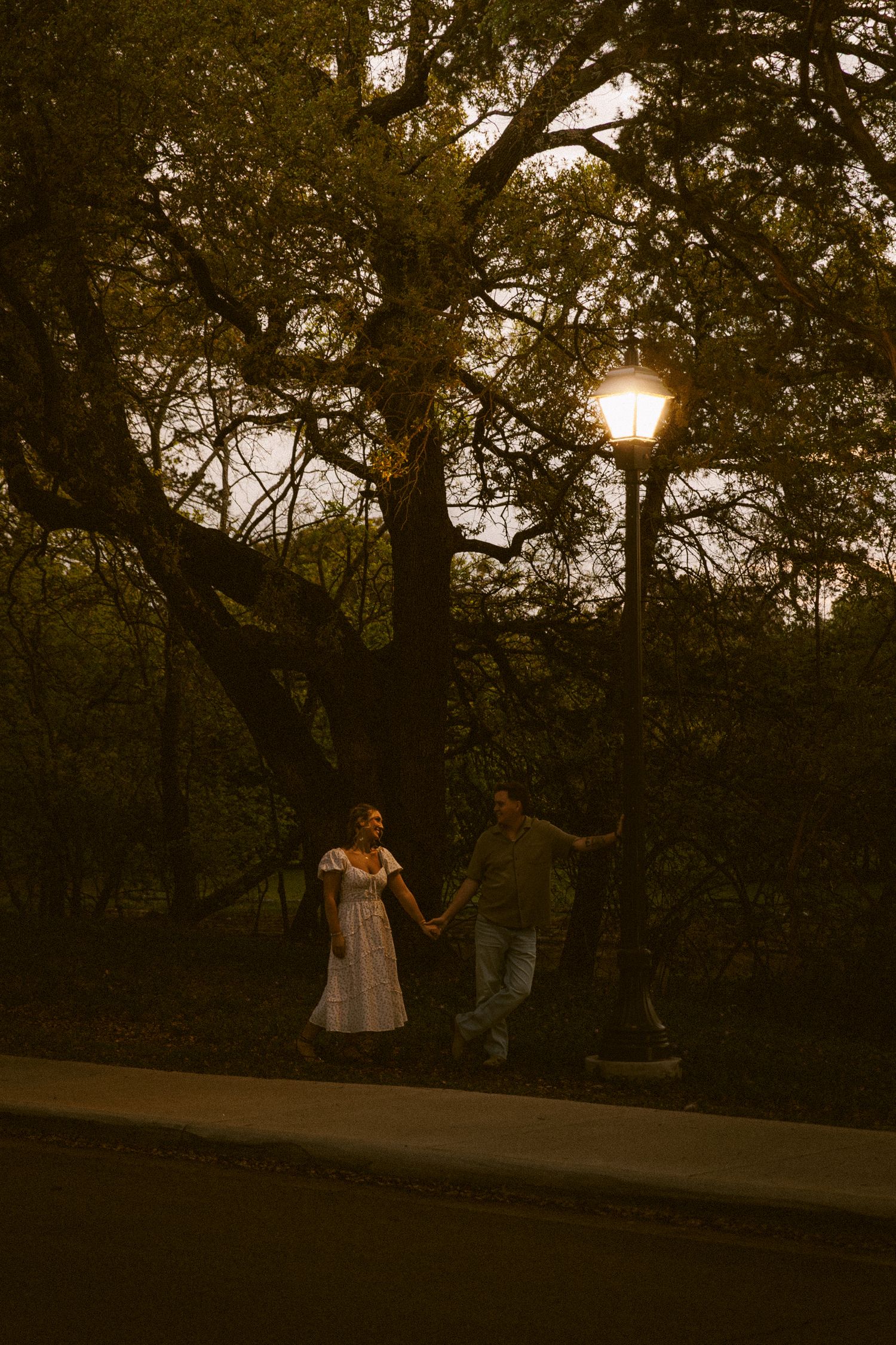 Couple walking hand in hand under a glowing street lamp among tall trees at twilight.