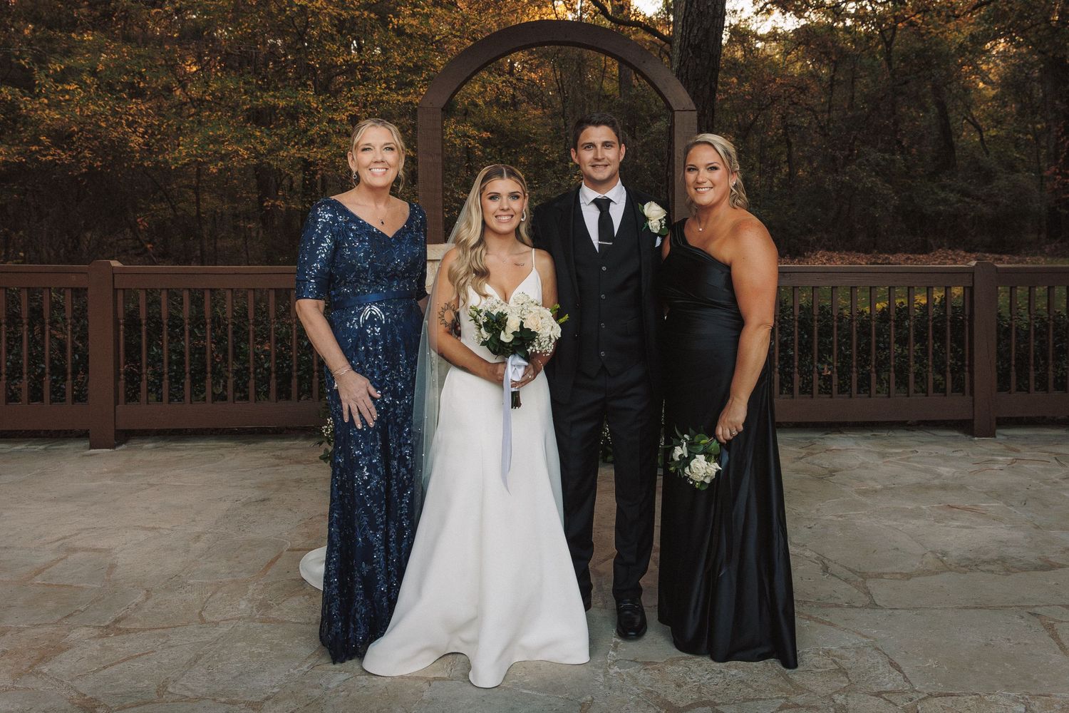 Wedding party of four posing together outdoors under a wooden arch at reception.