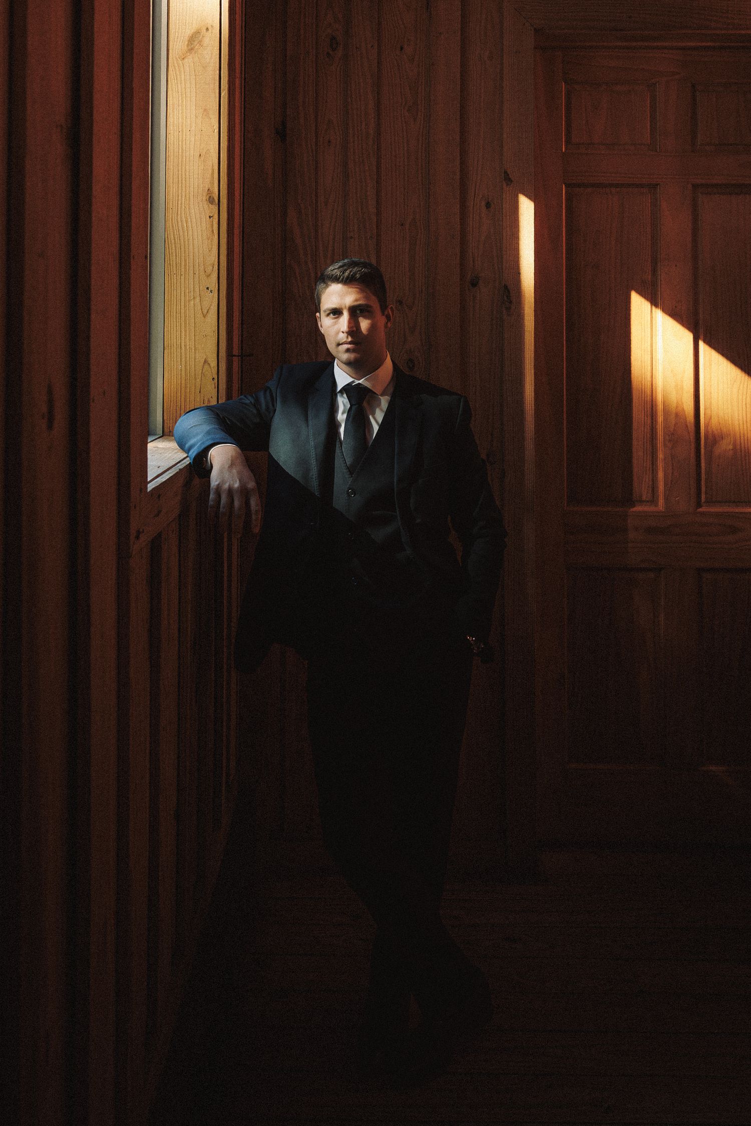 Handsome groom in black suit leaning against wooden door frame in dramatic warm lighting.