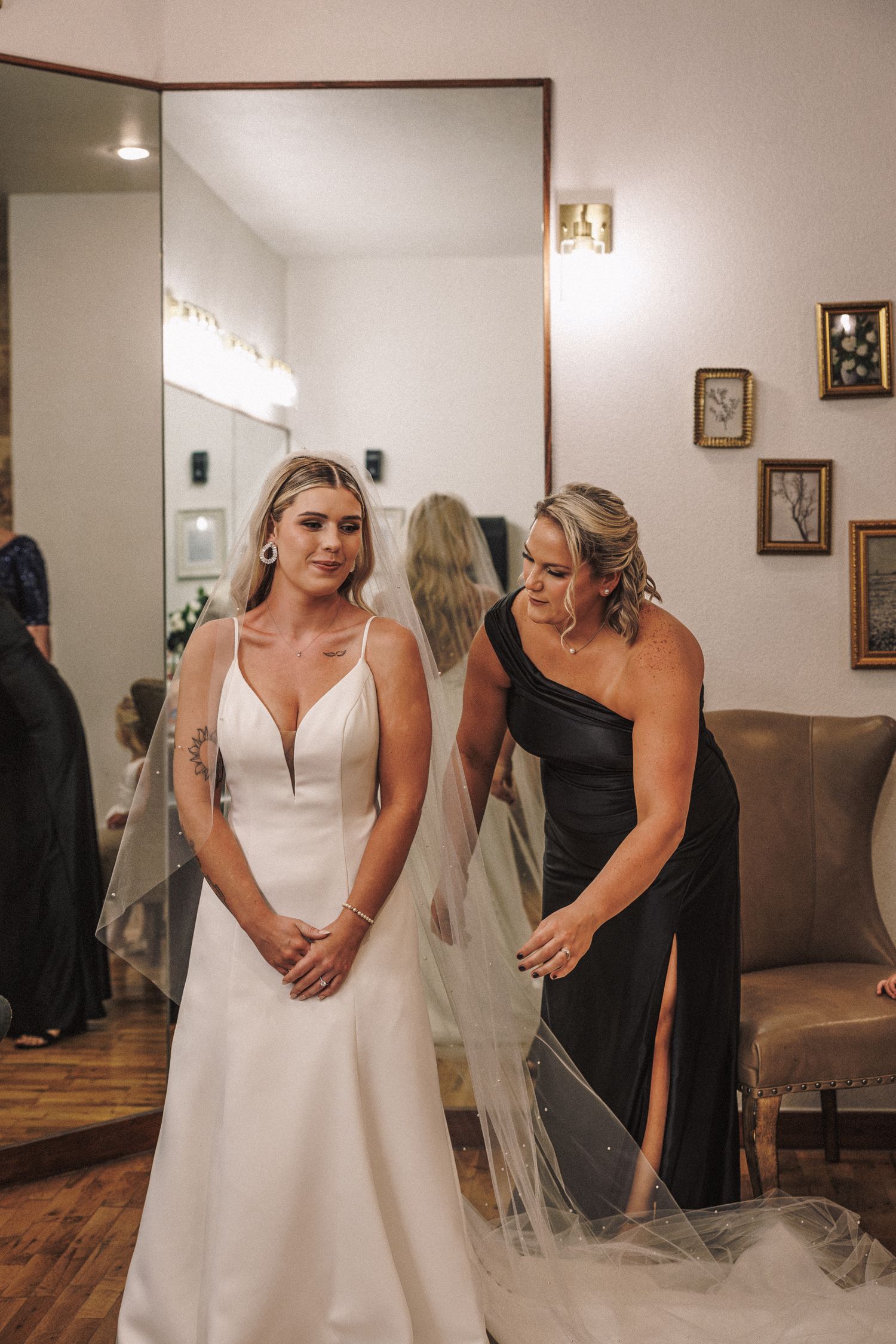 Bride in elegant white wedding gown with veil being adjusted by bridesmaid in black dress.