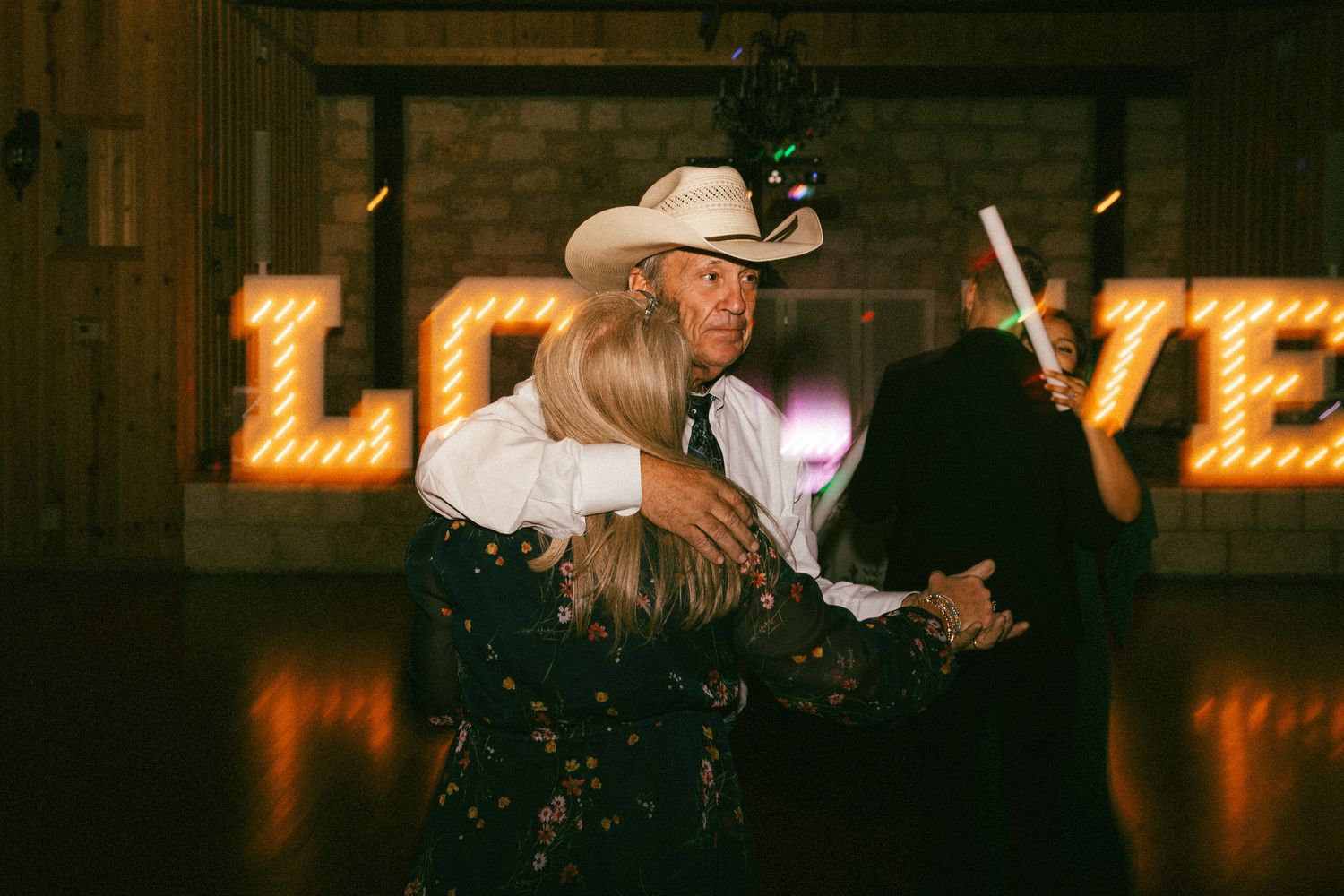 Three guests embrace on dance floor in front of illuminated LOVE marquee sign at wedding.