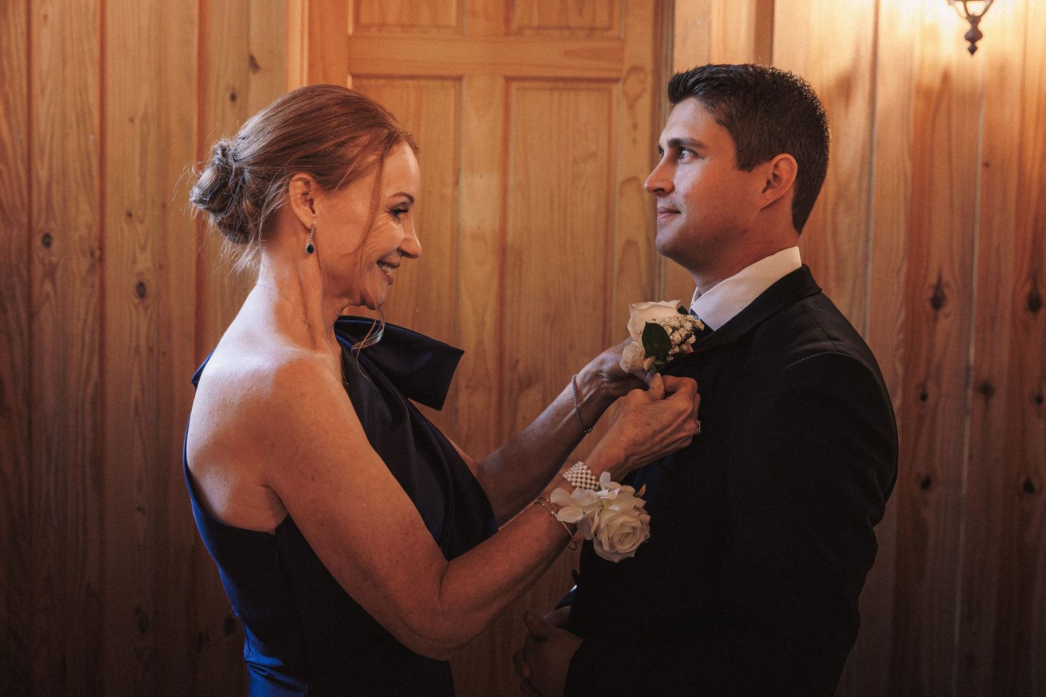 Woman in black dress pinning boutonniere on groom in tuxedo inside a warm wood-paneled room.