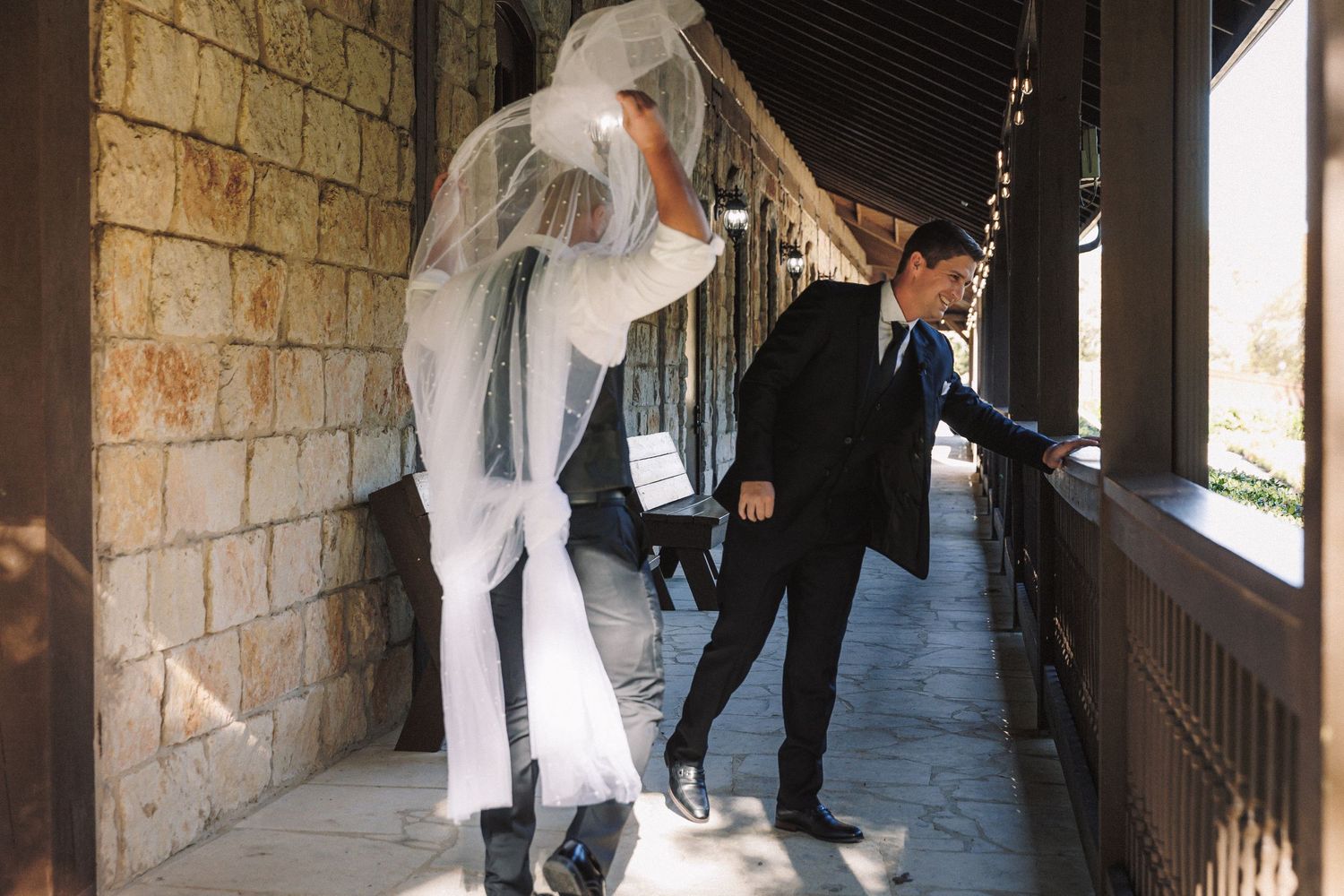 Bride in white gown and veil walking through a rustic stone corridor with groom nearby.
