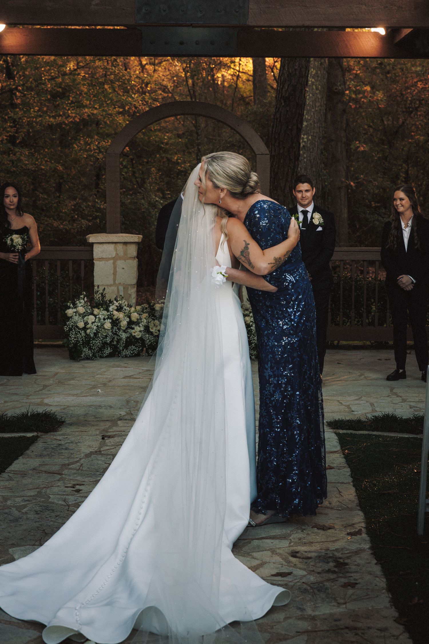Bride in white gown and veil embraces woman in blue sequin dress during outdoor wedding ceremony.