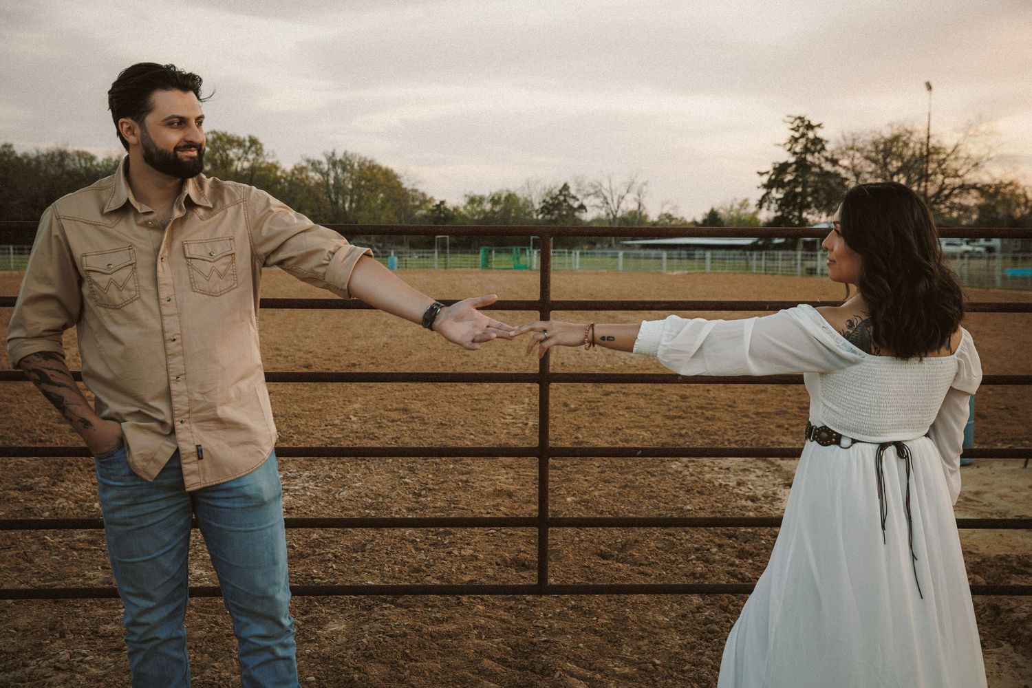 Couple reaching toward each other by a metal fence on a horse ranch.