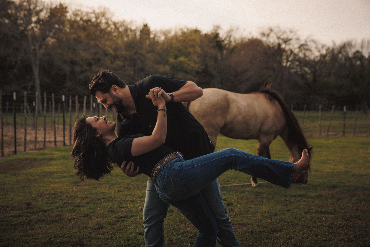 A couple playfully dancing in a green field with a horse grazing in the background at dusk.