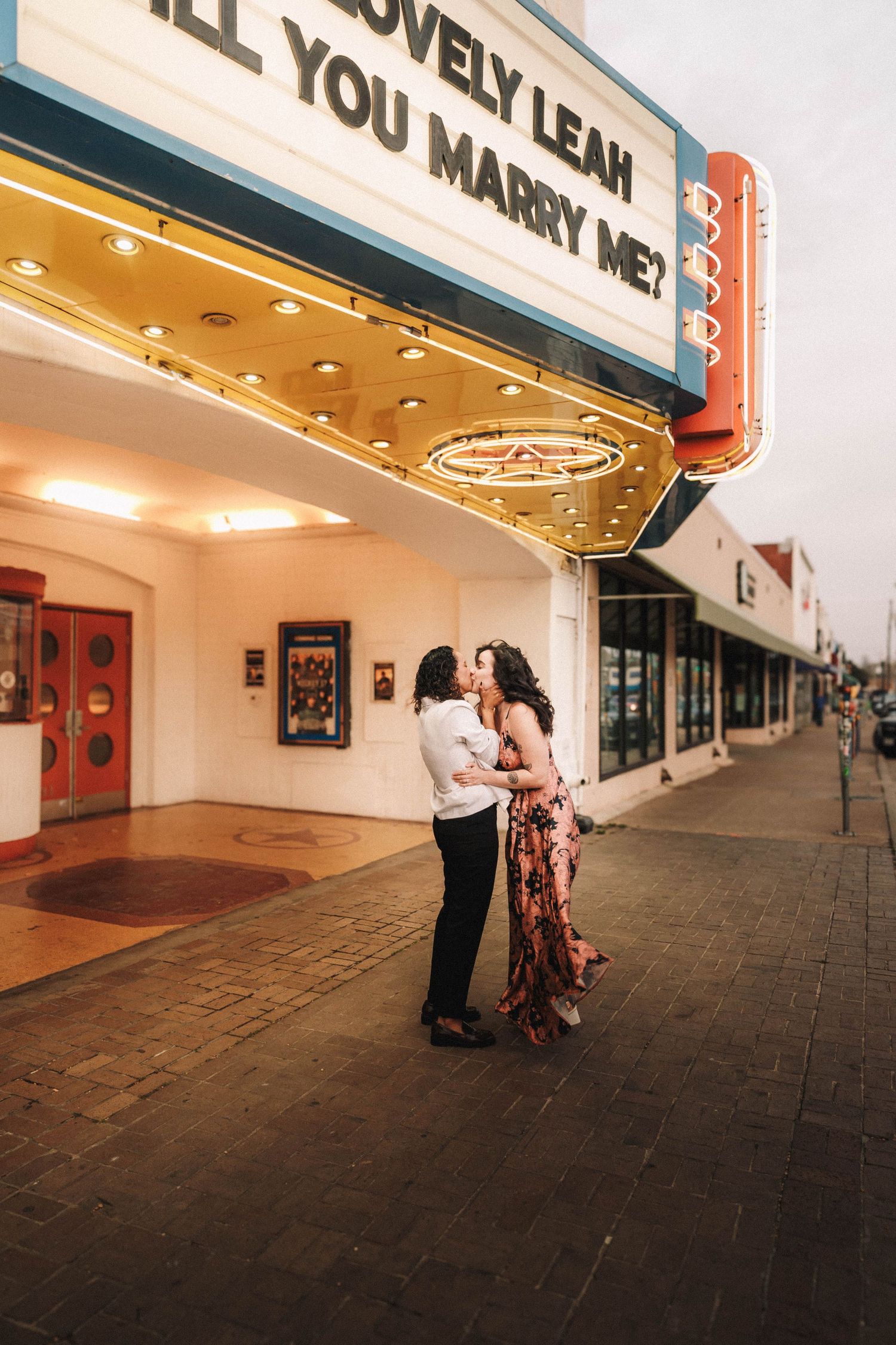 Couple kissing under glowing theater marquee displaying a marriage proposal for Leah on a city sidewalk at dusk.