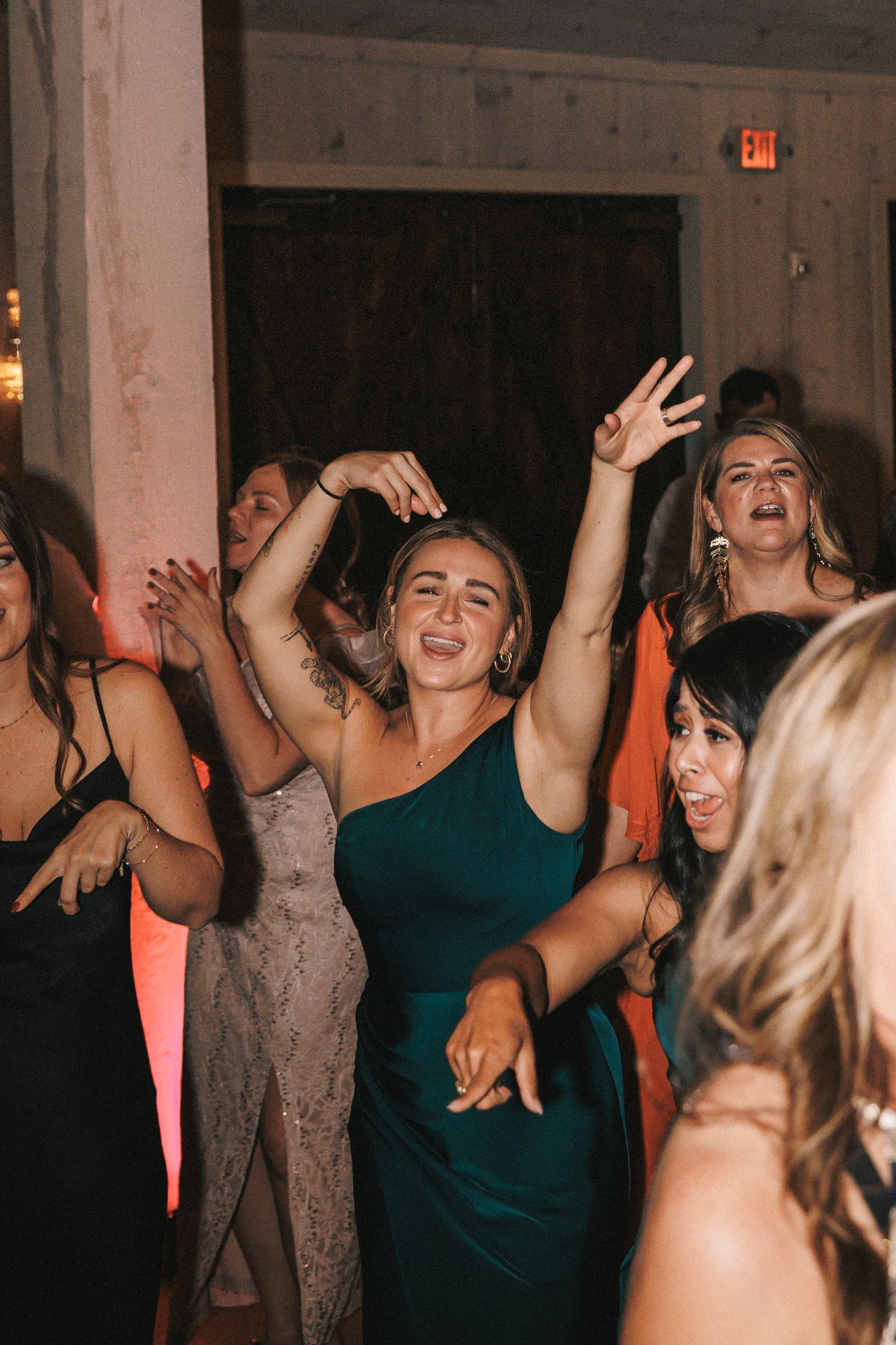 Bridesmaid in emerald green dress dances joyfully with hands raised at wedding reception.