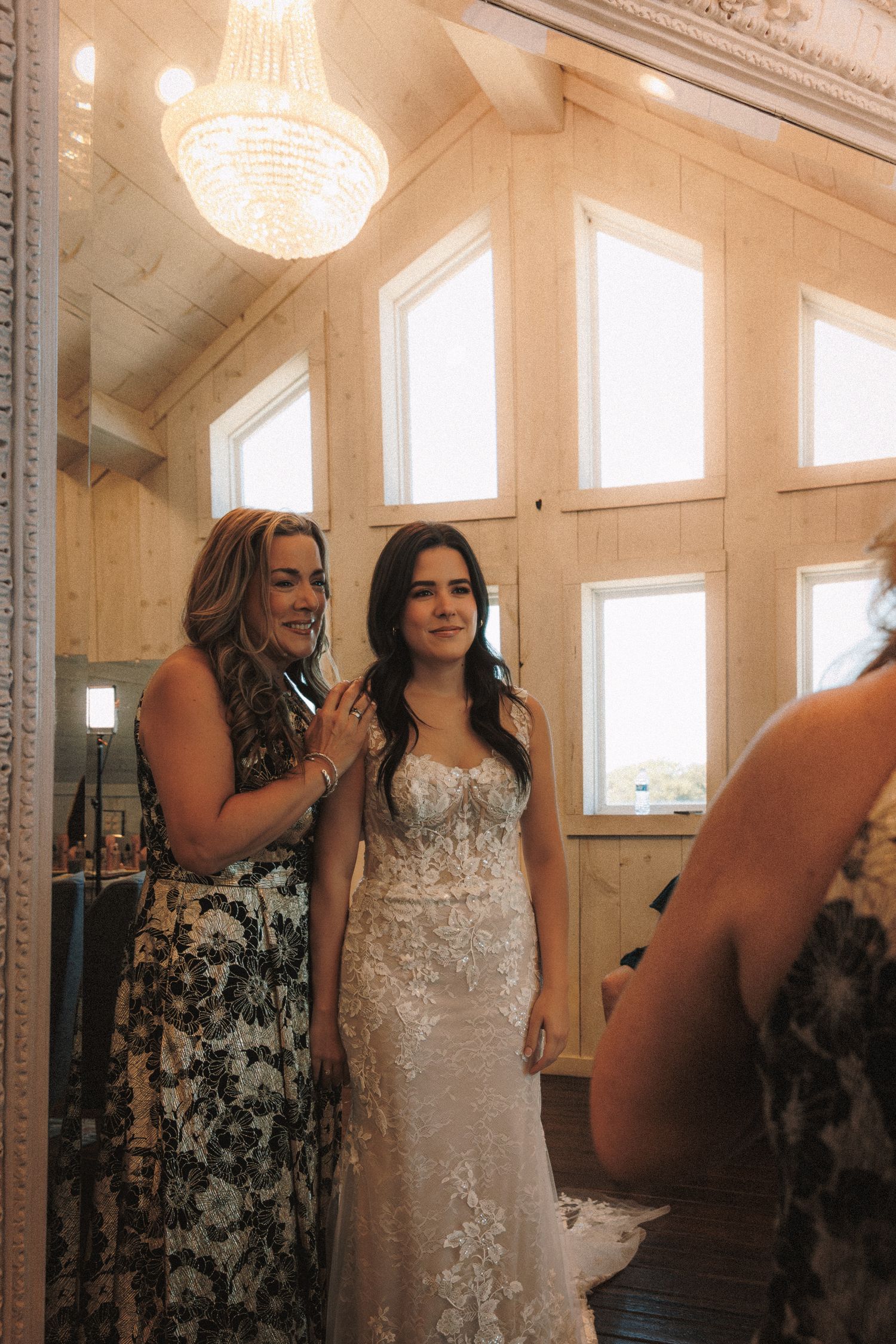 Bride in lace gown smiles with bridesmaid in floral dress inside elegant wood-paneled bridal suite with chandelier.