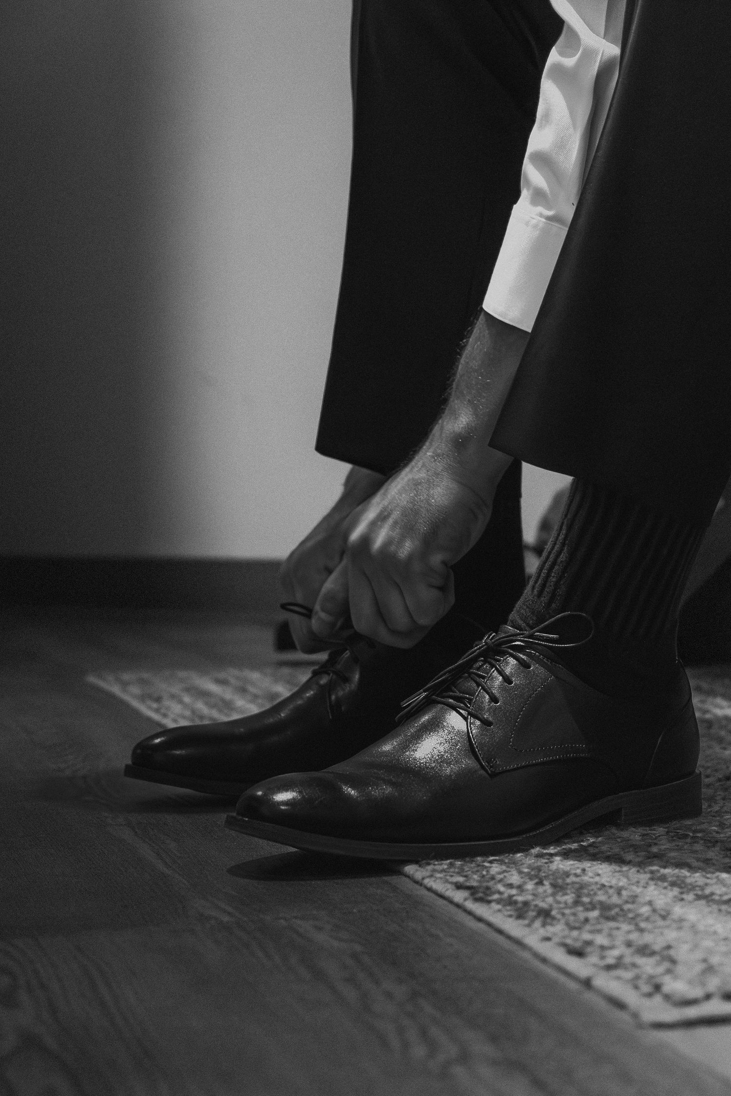 Groom tying laces on classic black leather dress shoes in black and white photo.