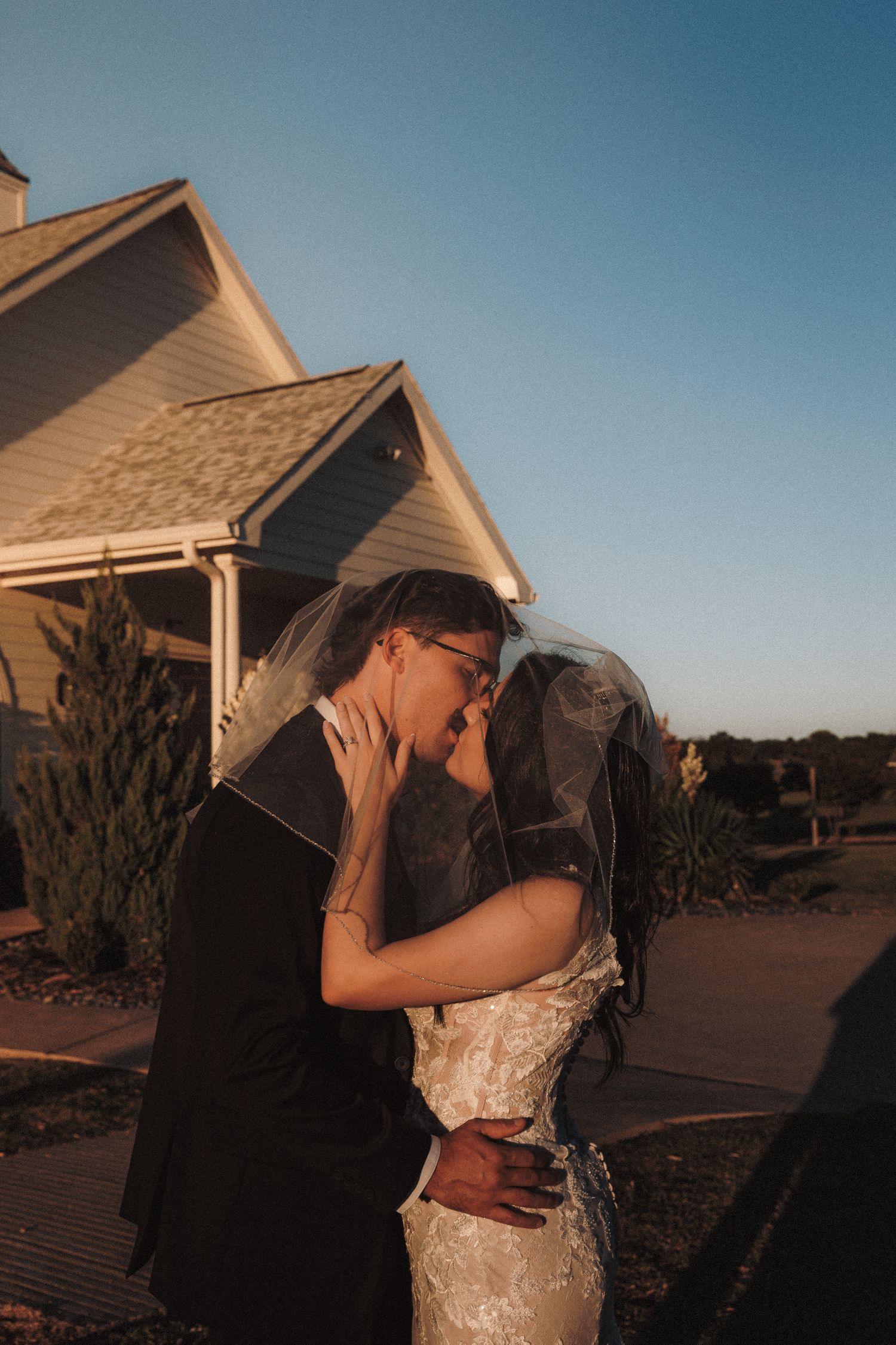 Bride and groom kiss under veil at golden hour outside white chapel after wedding ceremony.