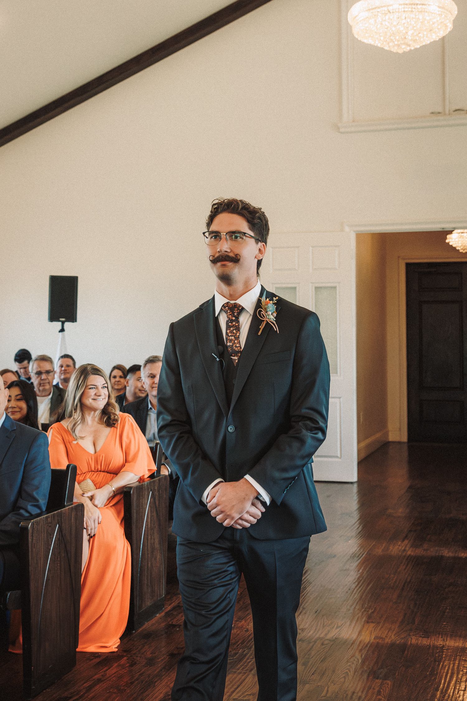 Groom in dark suit with floral tie and boutonniere waits at altar as guests fill chapel pews.