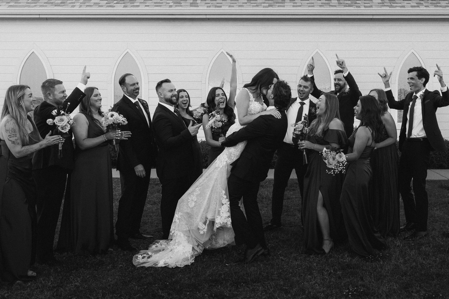 Black and white photo of wedding party cheering as groom dips bride outside a white chapel.