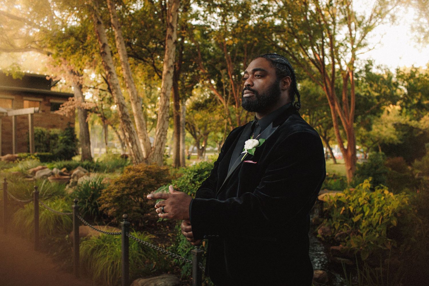 Groom in black velvet tuxedo with white boutonniere stands thoughtfully in a sunlit garden.
