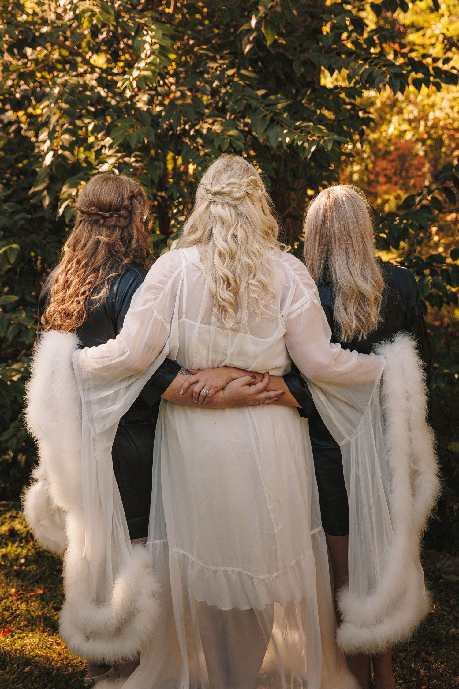 Bride in white sheer gown with two bridesmaids in black jackets and white fur wraps standing in autumn garden.