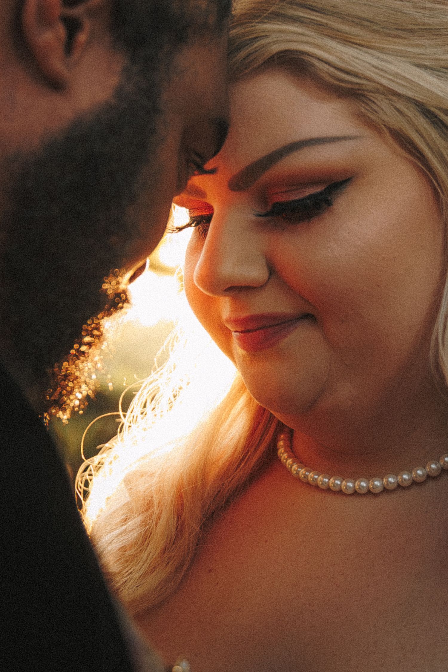 Bride wearing pearl necklace shares a close romantic moment with groom in golden sunset light.