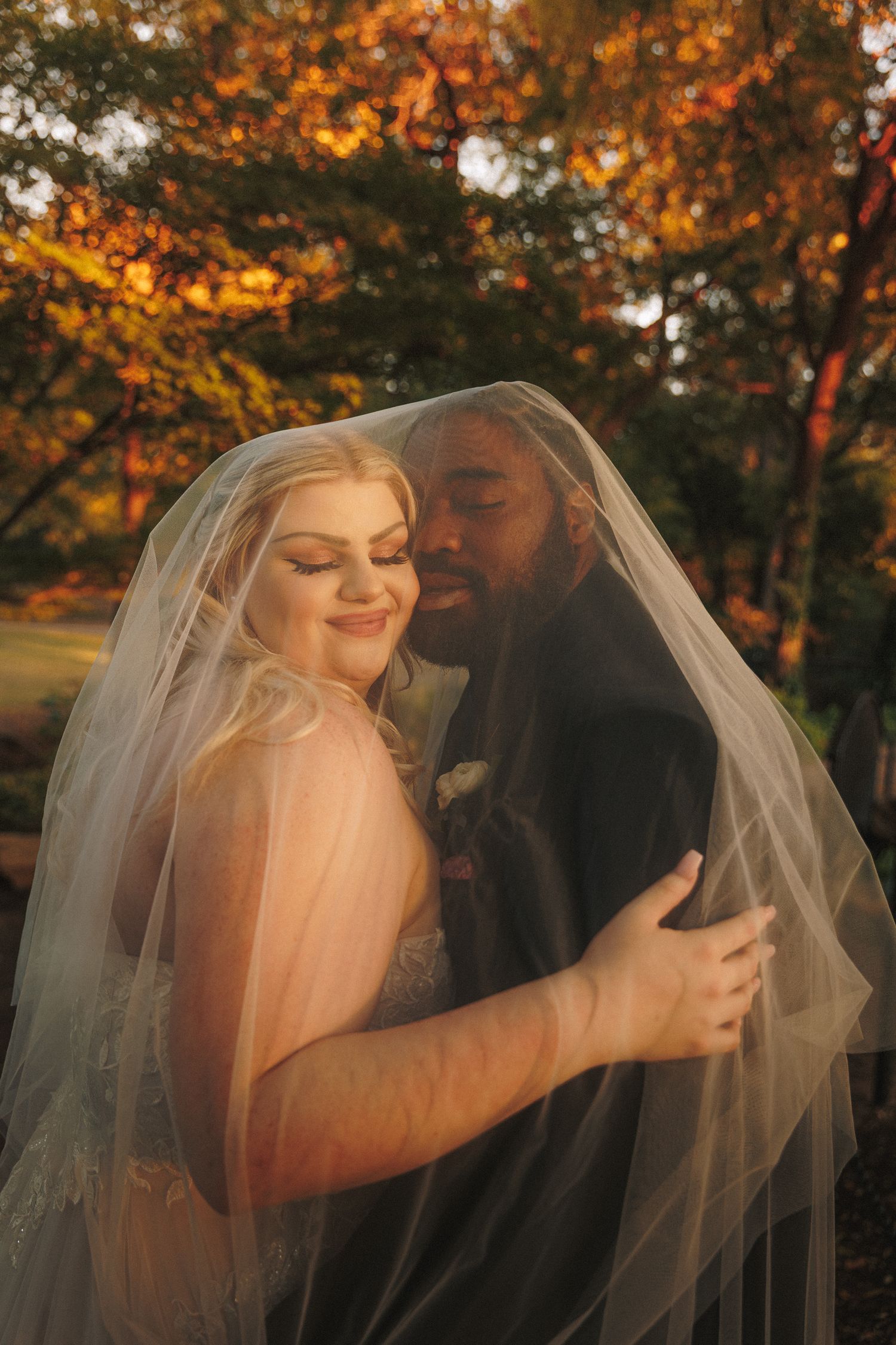 Bride and groom embrace under wedding veil surrounded by vibrant autumn foliage at sunset.