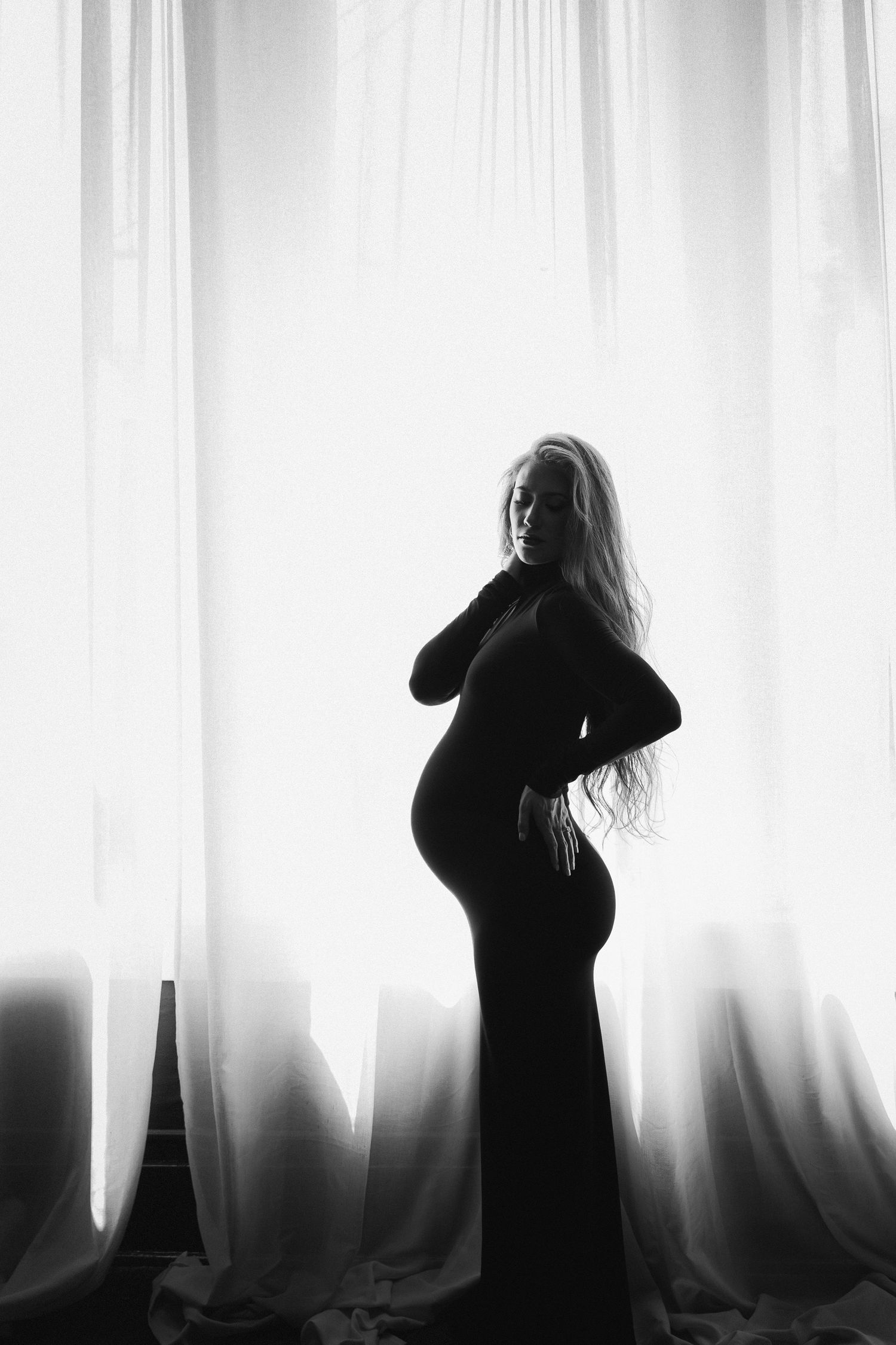 A silhouette of an expectant mother in a long dress stands gracefully against backlit white curtains in black and white.