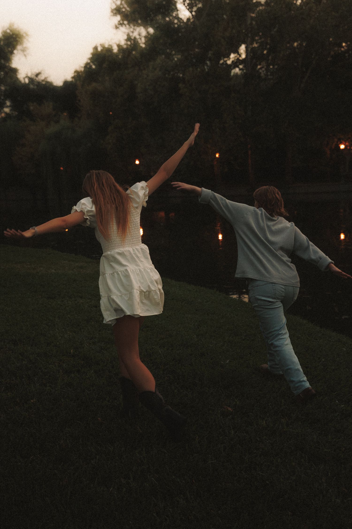 Two people dance together on a grassy hill at dusk with string lights glowing in the background.