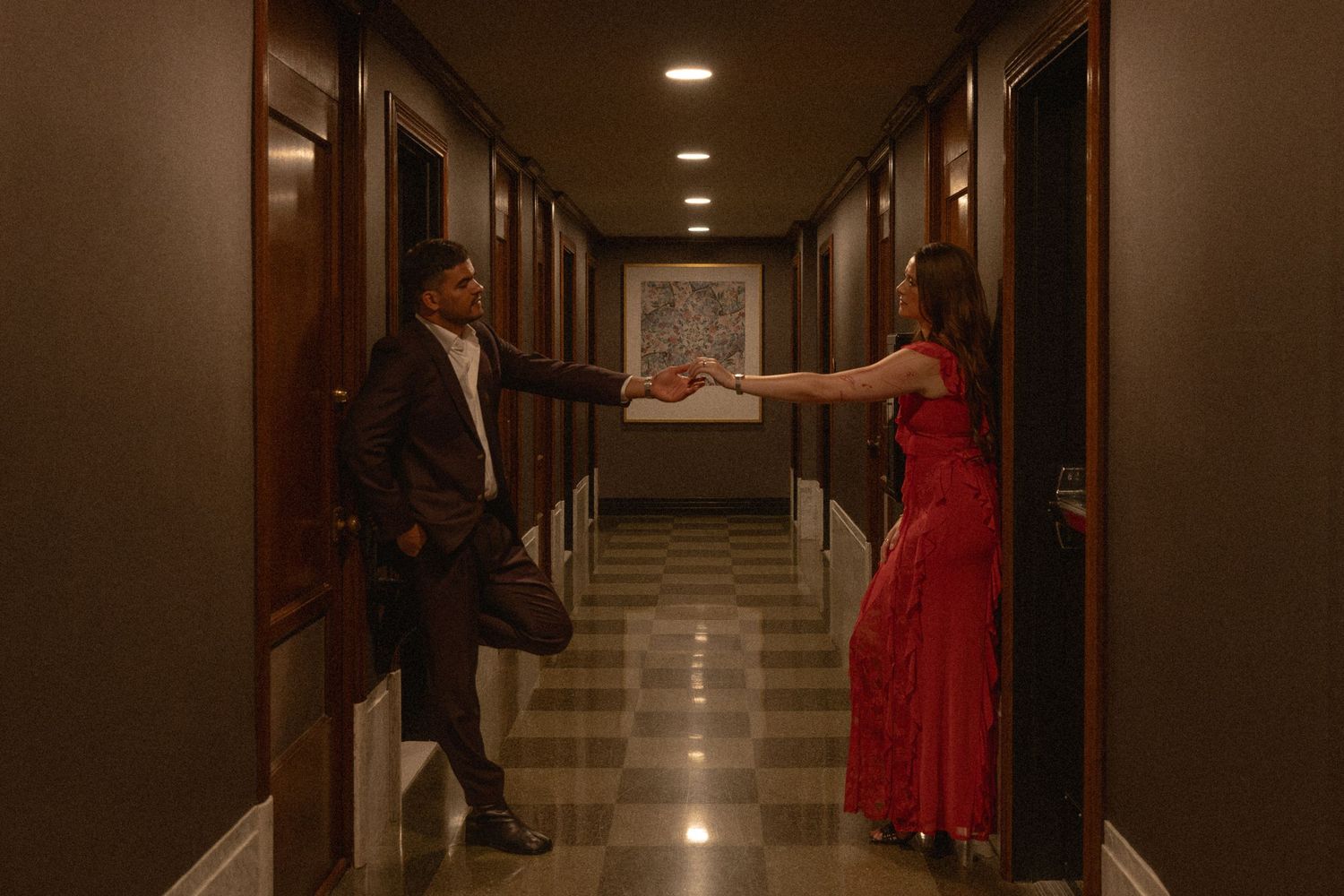 A man and woman in formal attire reach toward each other in a dimly lit hotel corridor.