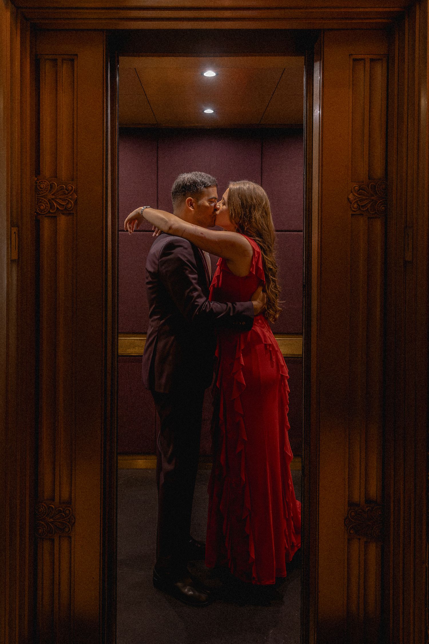 Couple sharing a romantic kiss in an elegant wooden elevator, woman in a flowing red dress embracing man in a dark suit.