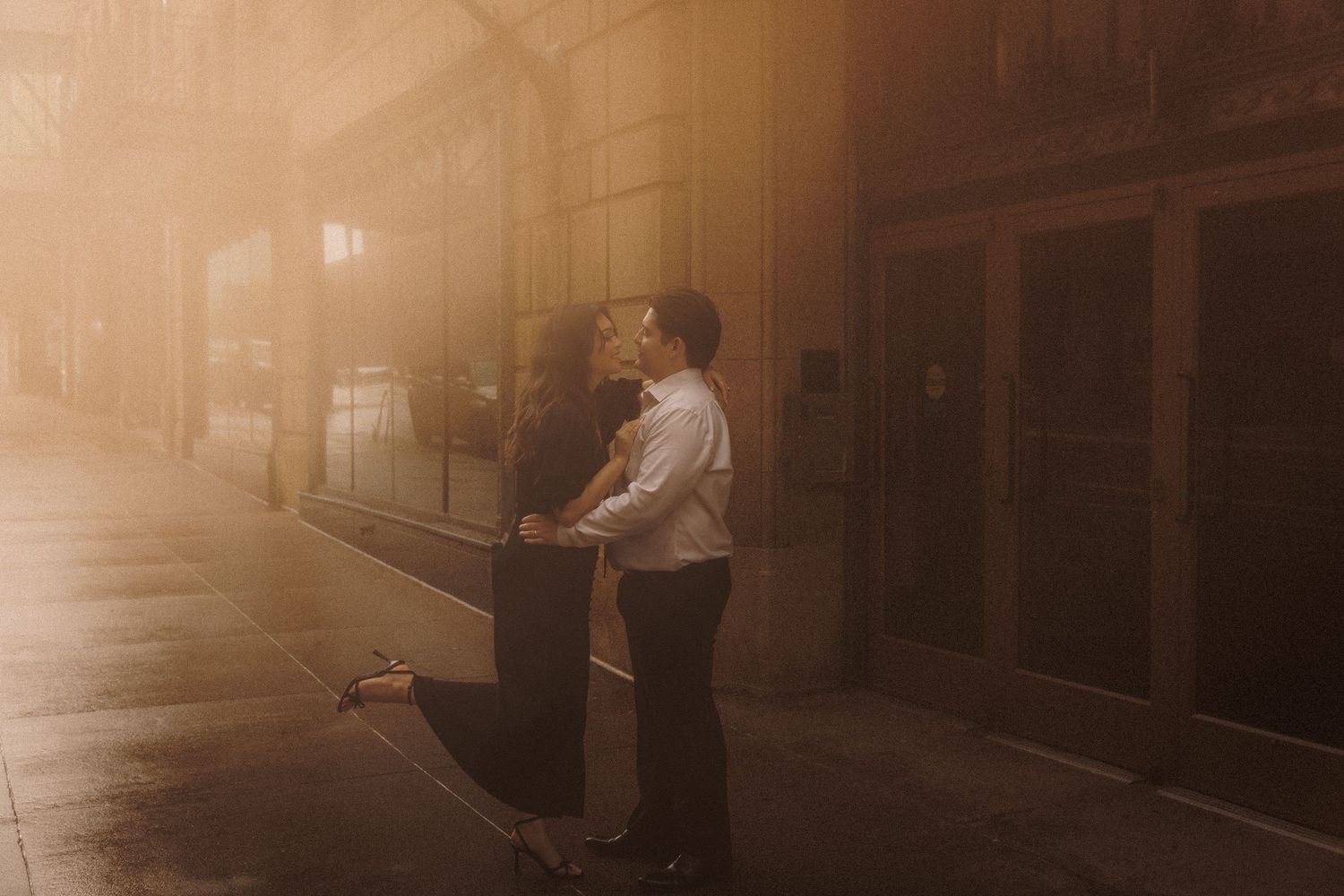 Couple sharing a romantic kiss on a misty urban sidewalk bathed in warm golden sunset light.