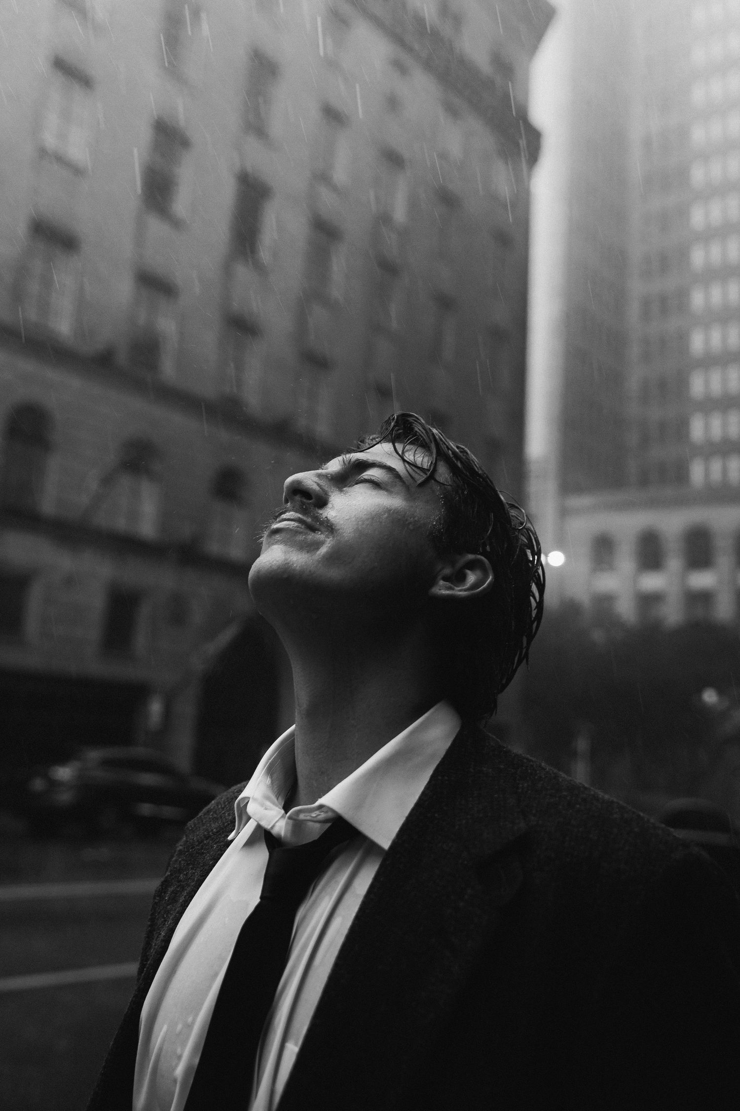 Black and white cityscape photo of person in business attire looking up at sky between tall urban buildings.