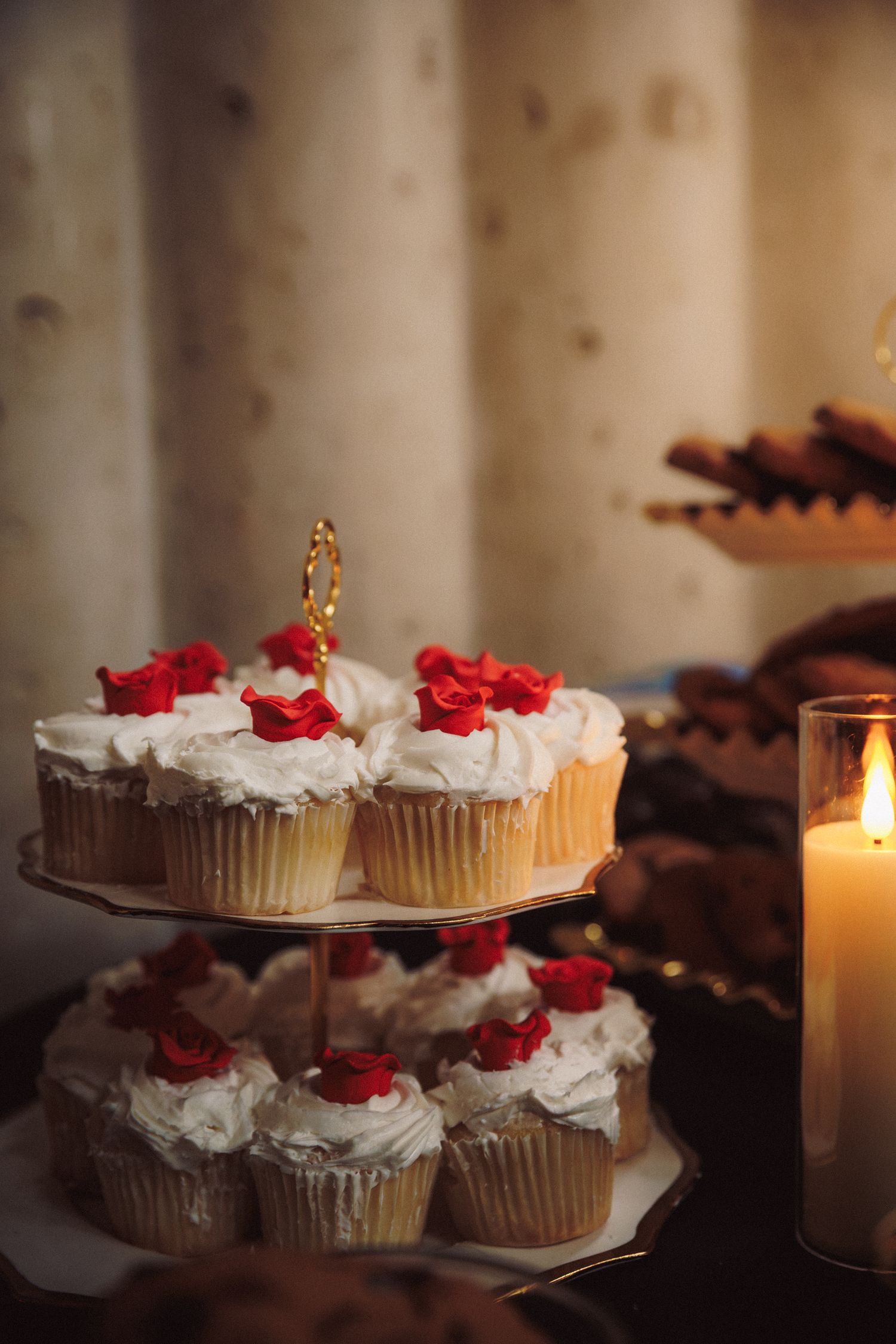 Elegant two-tier cupcake stand with white frosting and red rose decorations beside a glowing candle.