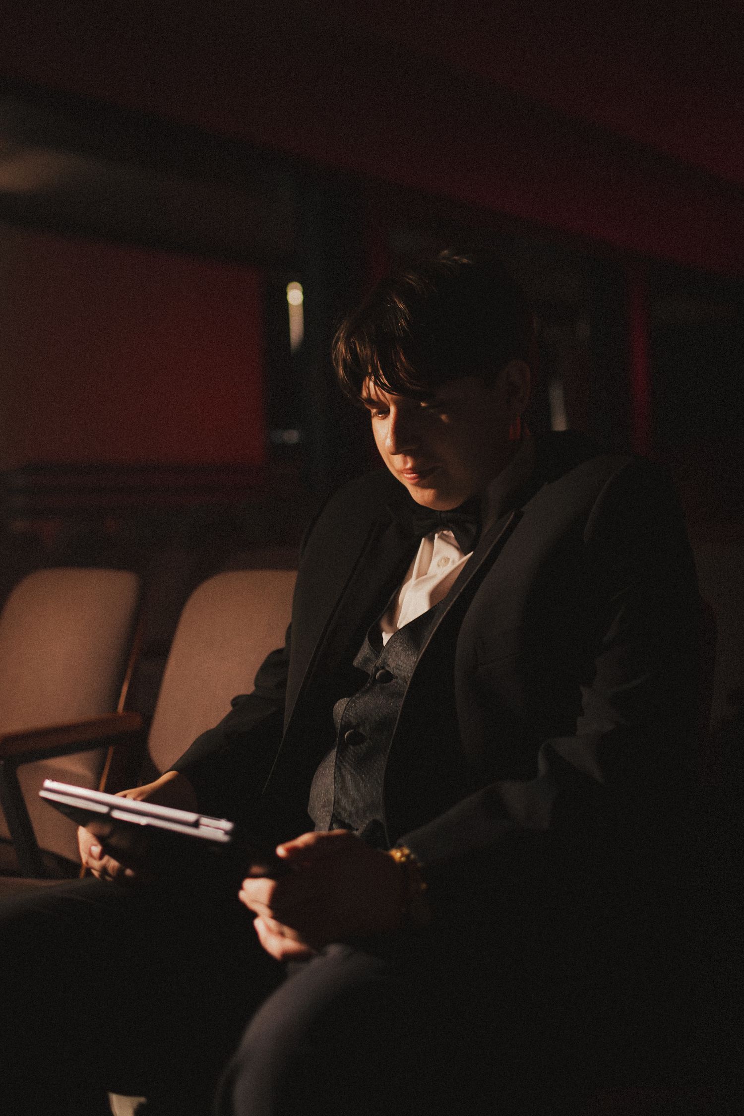 Groom in black suit reads notes in dimly lit theater seats before wedding ceremony.