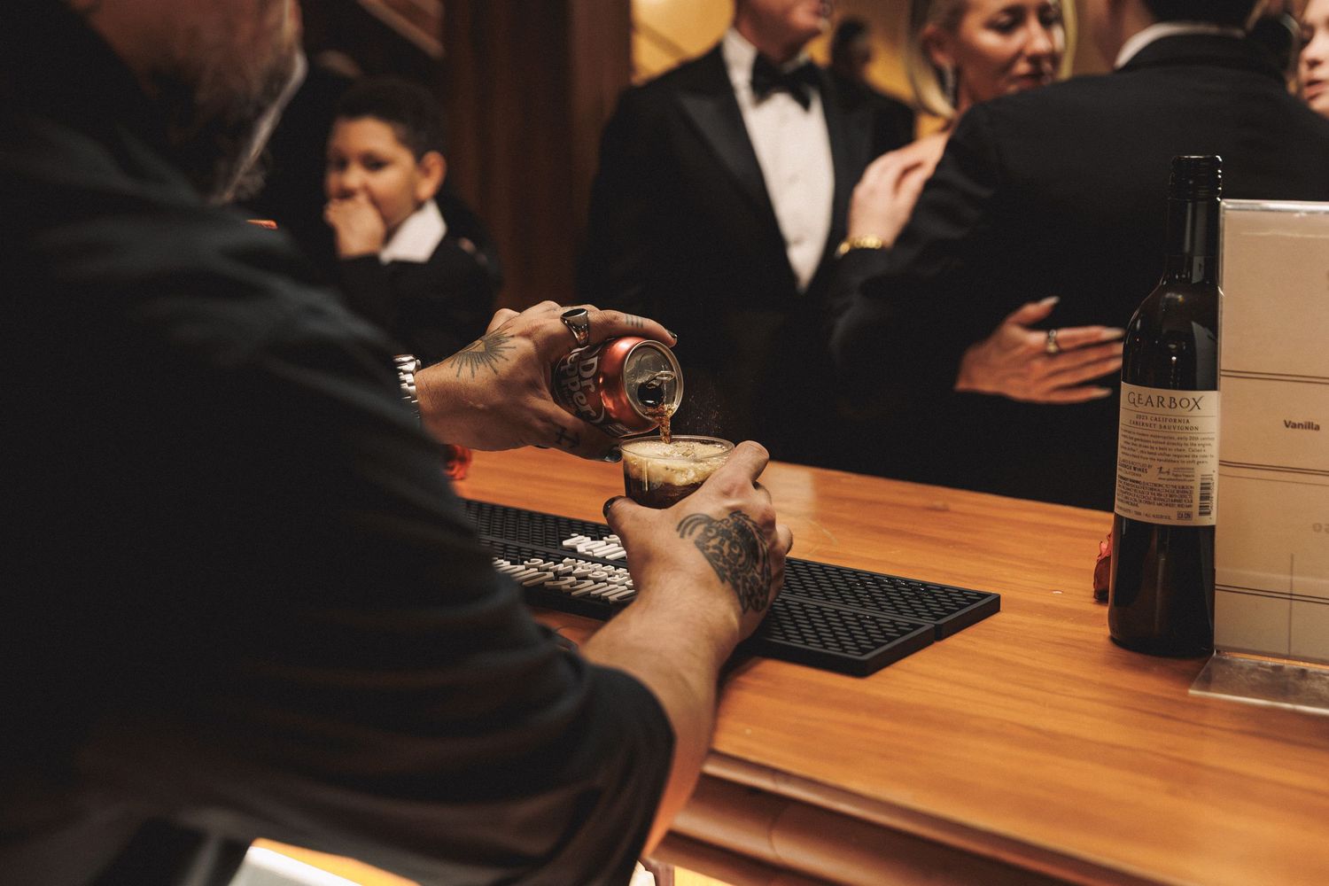Tattooed bartender pouring drink at wooden bar with guests in formal wear reflected in mirror.