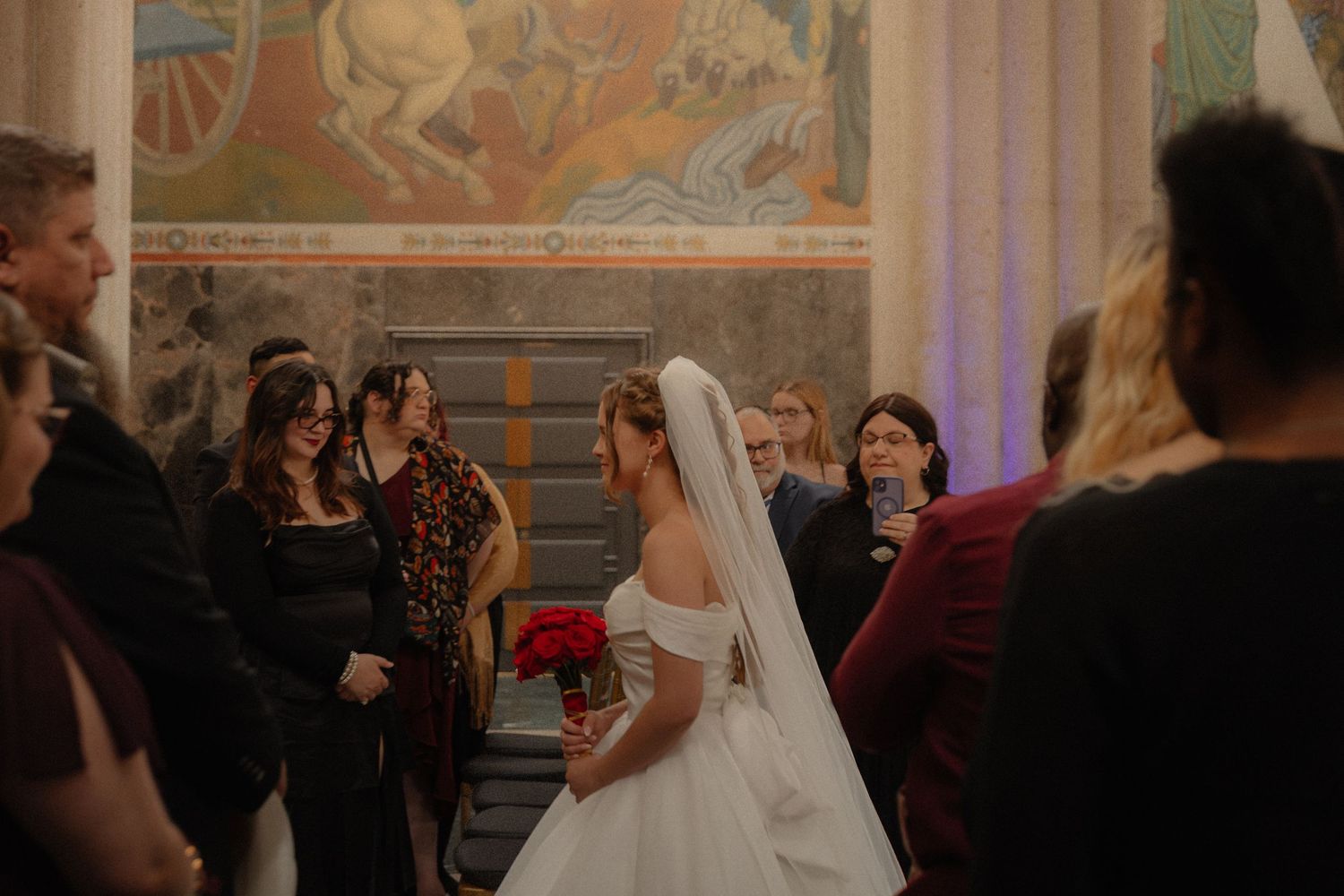 Bride in white ballgown with red bouquet walking down aisle past wedding guests.